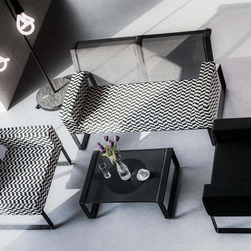 Overhead view of a modern living space featuring an Egoe Moja sofa in black and white pattern, a sleek black sofa, and modular collection elements. A glass-topped coffee table with flowers, pitcher, and cup rests near a floor lamp with looped bulbs on the light-colored floor.