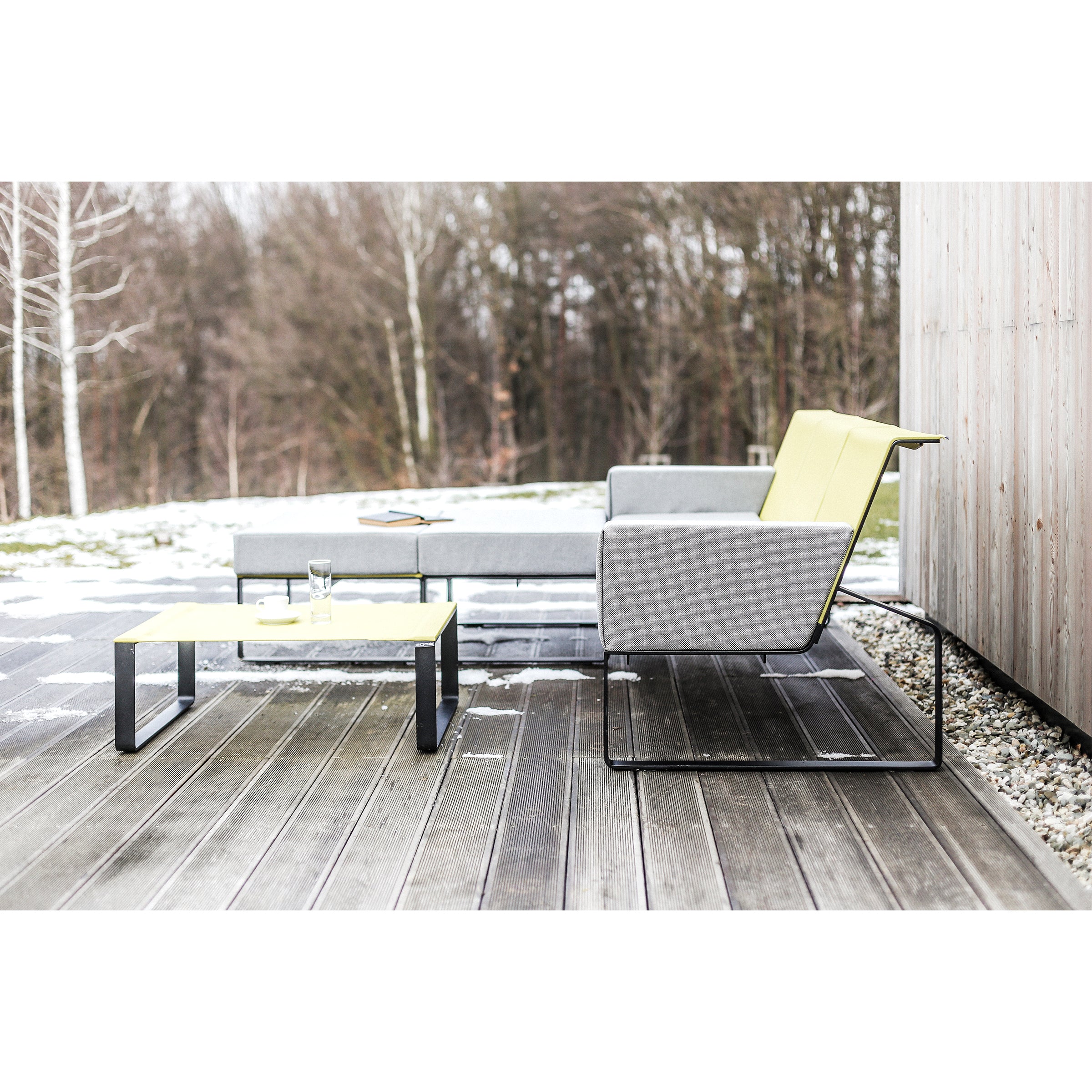 A modern outdoor patio with Egoes Moja Sofa in minimalist gray and yellow sits on a wooden deck. A low table features a glass and plate, while sparse trees and light snow enhance the cozy feel of this inviting seating area.