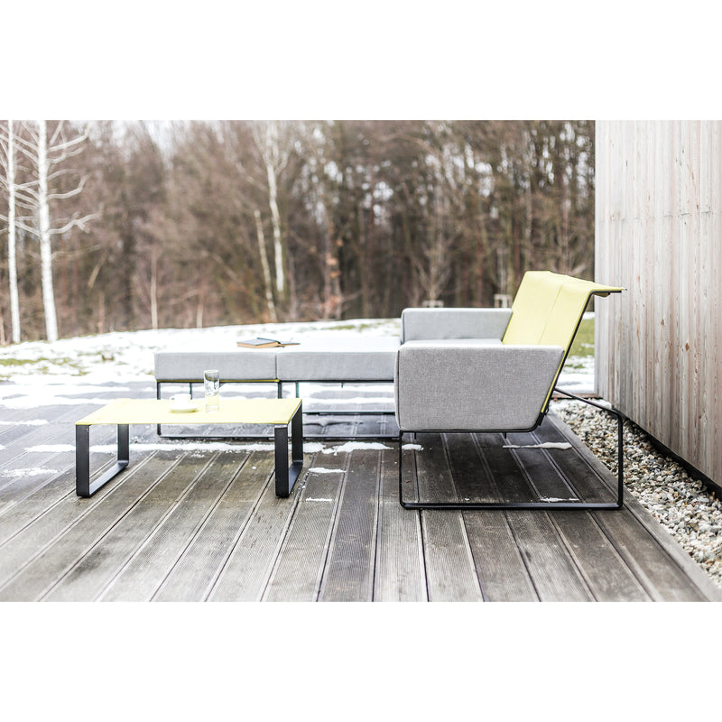 A modern outdoor patio with Egoes Moja Sofa in minimalist gray and yellow sits on a wooden deck. A low table features a glass and plate, while sparse trees and light snow enhance the cozy feel of this inviting seating area.