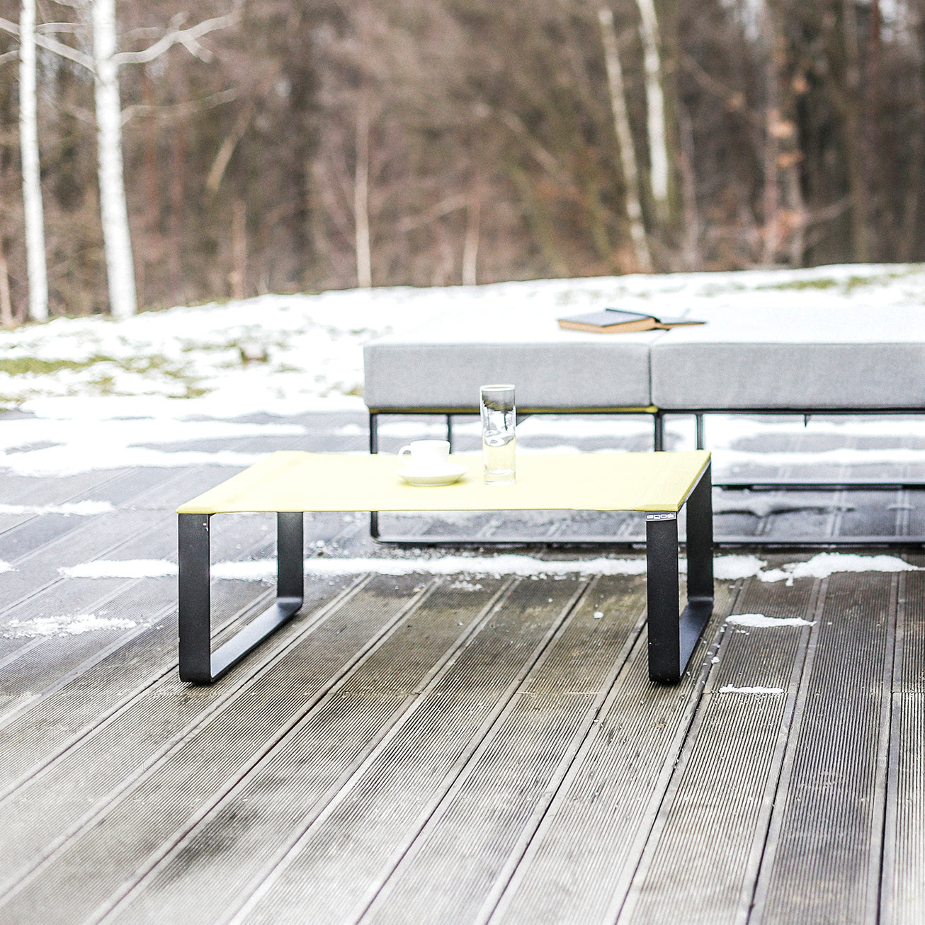 The Moja Coffee Table by Egoe, featuring a glass and bowl, sits on a snowy wooden deck. Behind it, a gray outdoor sofa from a sleek modular collection has a closed book atop. Bare trees form the backdrop, adding to this serene outdoor seating space.