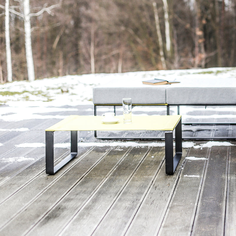 The Moja Coffee Table by Egoe, featuring a glass and bowl, sits on a snowy wooden deck. Behind it, a gray outdoor sofa from a sleek modular collection has a closed book atop. Bare trees form the backdrop, adding to this serene outdoor seating space.