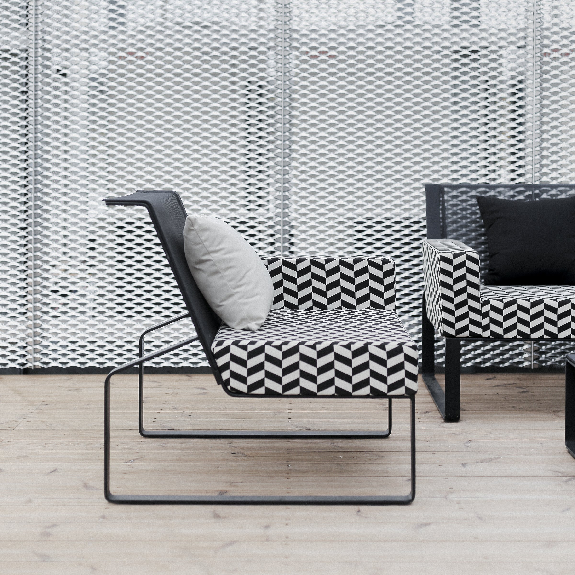 A modern outdoor seating area features striking Moja Lounge Chairs by Egoe, with black metal frames, Batyline outdoor mesh, and geometric black-and-white patterned cushions. One chair boasts a white cushion against a textured light-colored decorative wall, enhancing the serene atmosphere.