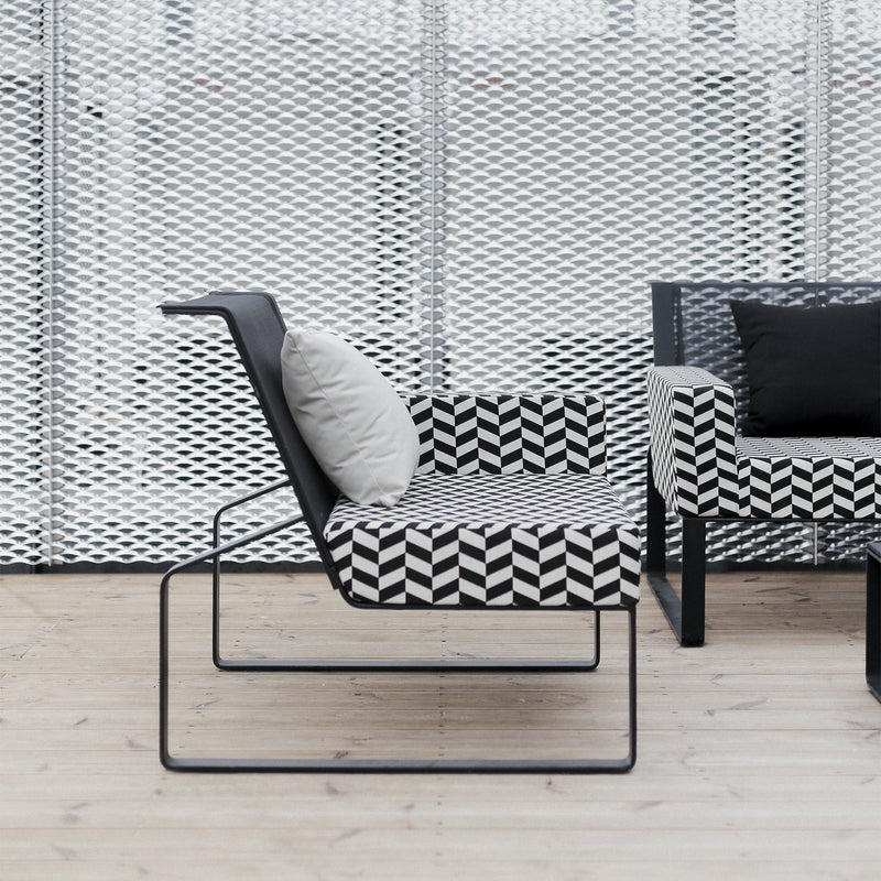 A modern outdoor seating area features striking Moja Lounge Chairs by Egoe, with black metal frames, Batyline outdoor mesh, and geometric black-and-white patterned cushions. One chair boasts a white cushion against a textured light-colored decorative wall, enhancing the serene atmosphere.