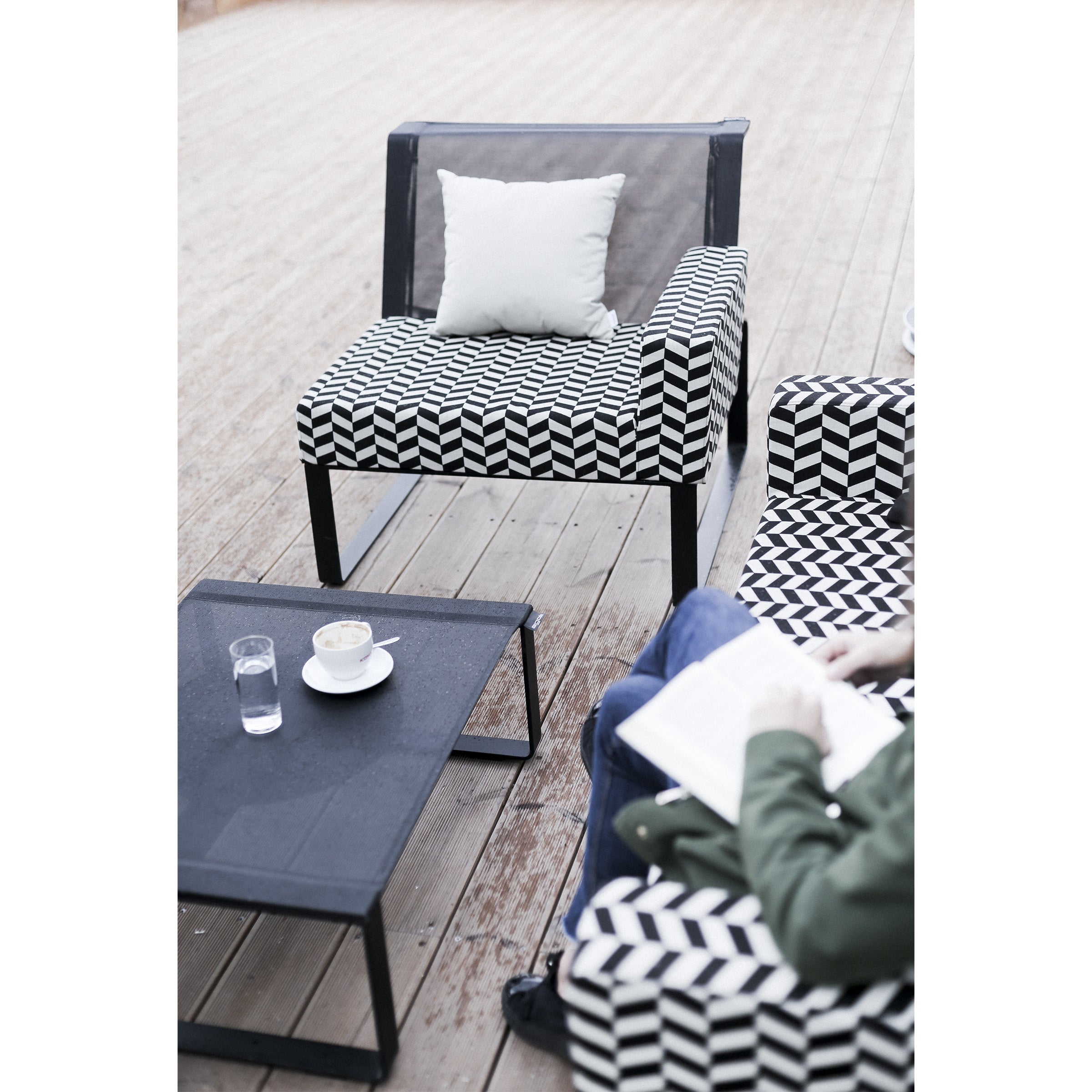 A person sits on the Moja Lounge Chair by Egoe, reading a book next to a black coffee table topped with a glass of water and a cup of cappuccino. This outdoor setup includes another empty chair with a white cushion, all set on a wooden deck featuring geometric patterns.