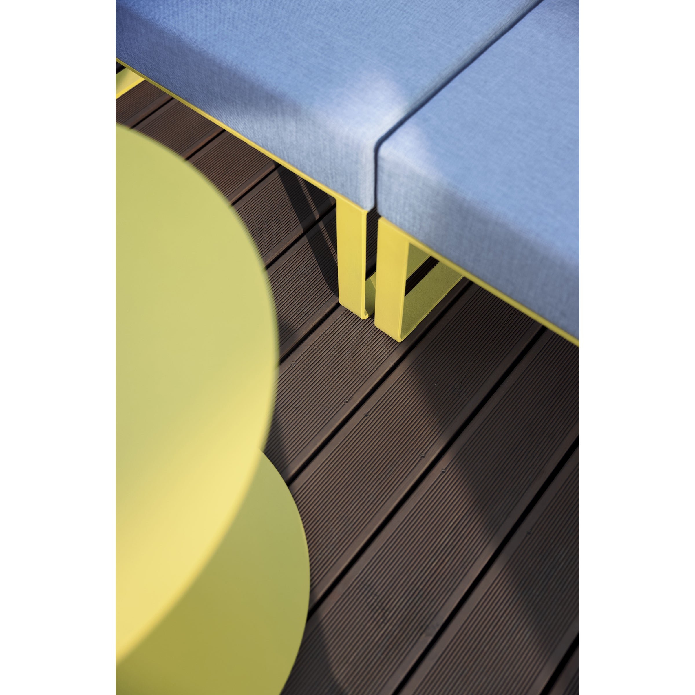 The Moja modular collection, featuring a curved light green table next to a blue-cushioned bench with yellow legs, crafted with galvanized steel by Egoe, adds vibrance to a modern outdoor setting as it contrasts against the dark wooden deck.