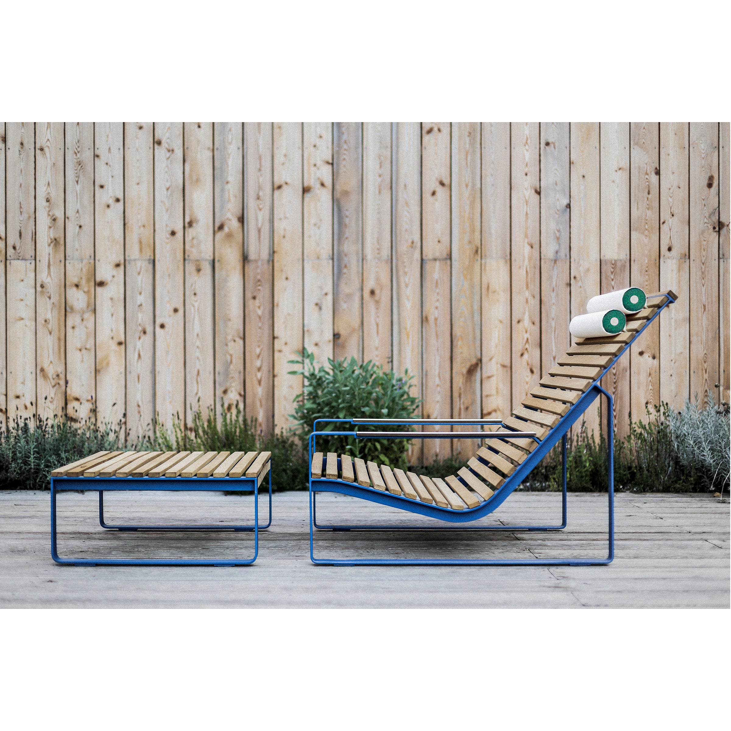 The Preva Lounger by Egoe exudes sophistication with its wooden design and blue metal frame, accompanied by a matching ottoman. Two green and white rolled towels enhance the setup against a rustic fence and lush plants on the wooden deck.
