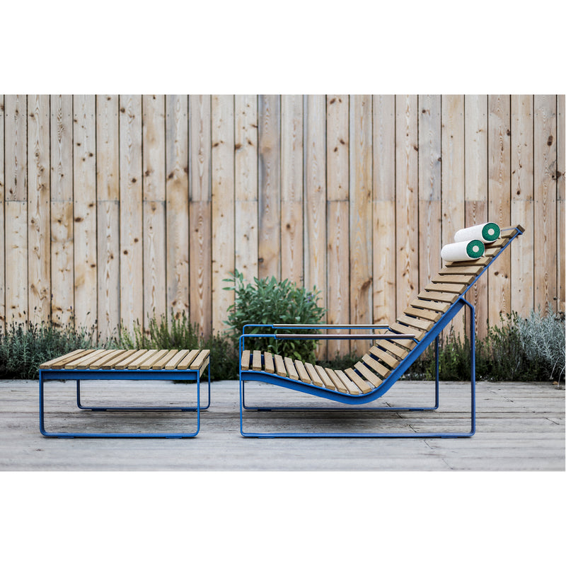 The Preva Ottoman by Egoe, a modern wooden lounge chair with a sleek metal frame, exudes sophisticated style against a wooden fence backdrop. A green-striped towel rests on the chair, while shrubs line the fence base, completing this high-quality furniture scene.