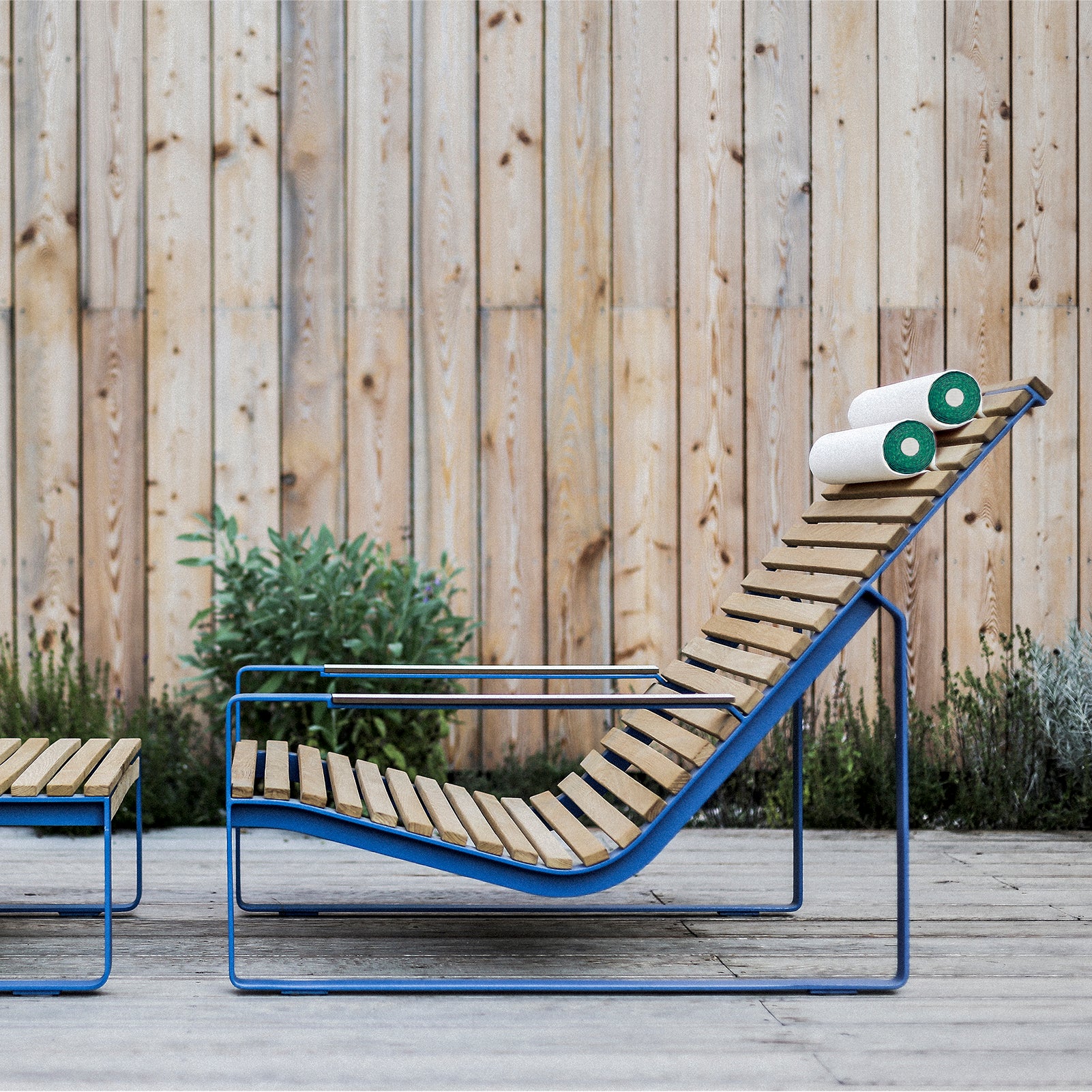 The Preva Lounger by Egoe, made of tropical wood with a blue metal frame, is set on a stone patio. It includes a minimalist side table and has two rolled white towels at the headrest, surrounded by a wooden fence and lush greenery.