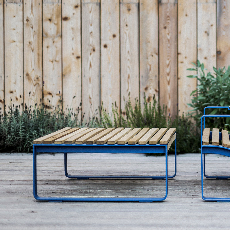 Two Preva Ottomans by Egoe, featuring wooden slats and blue metal frames, sit on a wooden deck. Paired with a wooden fence and green plants, this high-quality furniture creates a serene garden setting with sophisticated style.