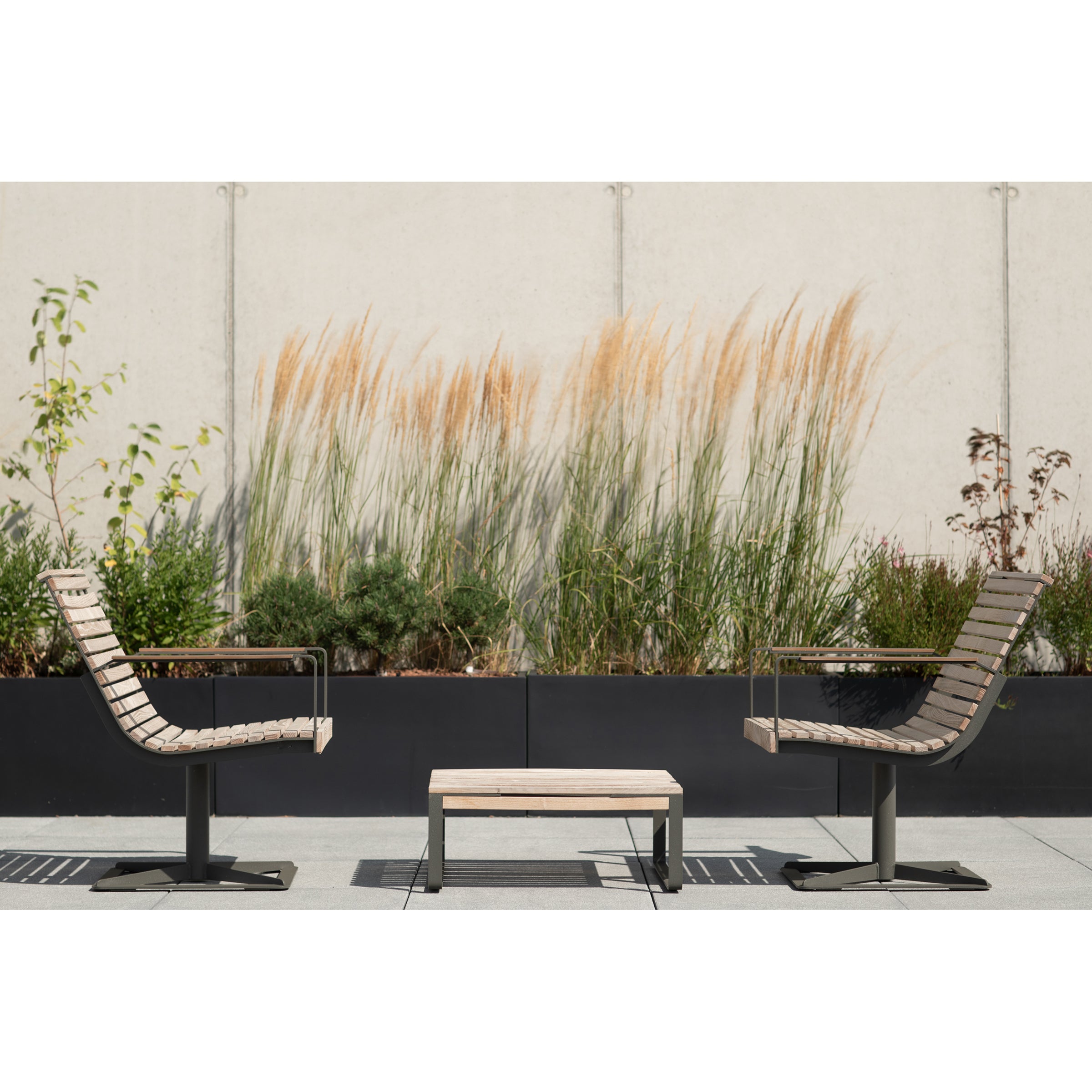 Two Preva Armchairs by Egoe flank a small table on the patio. They are backed by tall ornamental grasses and greenery in planters against a light concrete wall, creating a sophisticated and minimalistic escape.