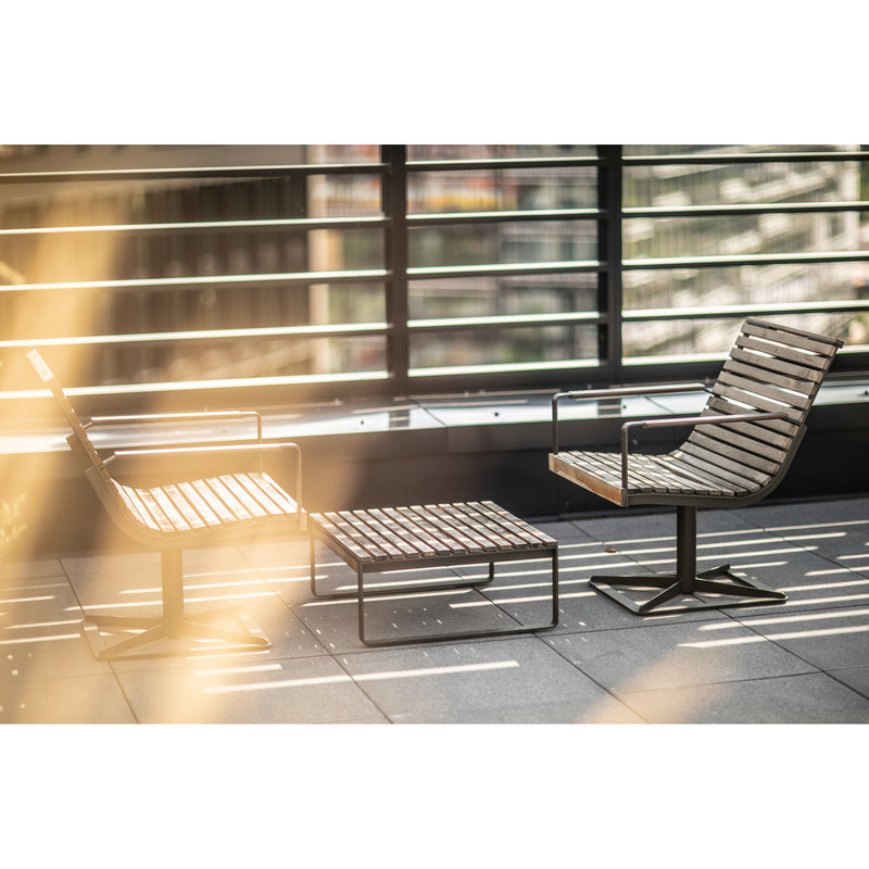 Two durable Preva Armchairs by Egoe and a matching table adorn a sunlit patio. The black metal railings partially obscure the city view, crafting a serene outdoor space. Soft sunlight casts warm reflections on the sophisticated furniture design and tiled floor.