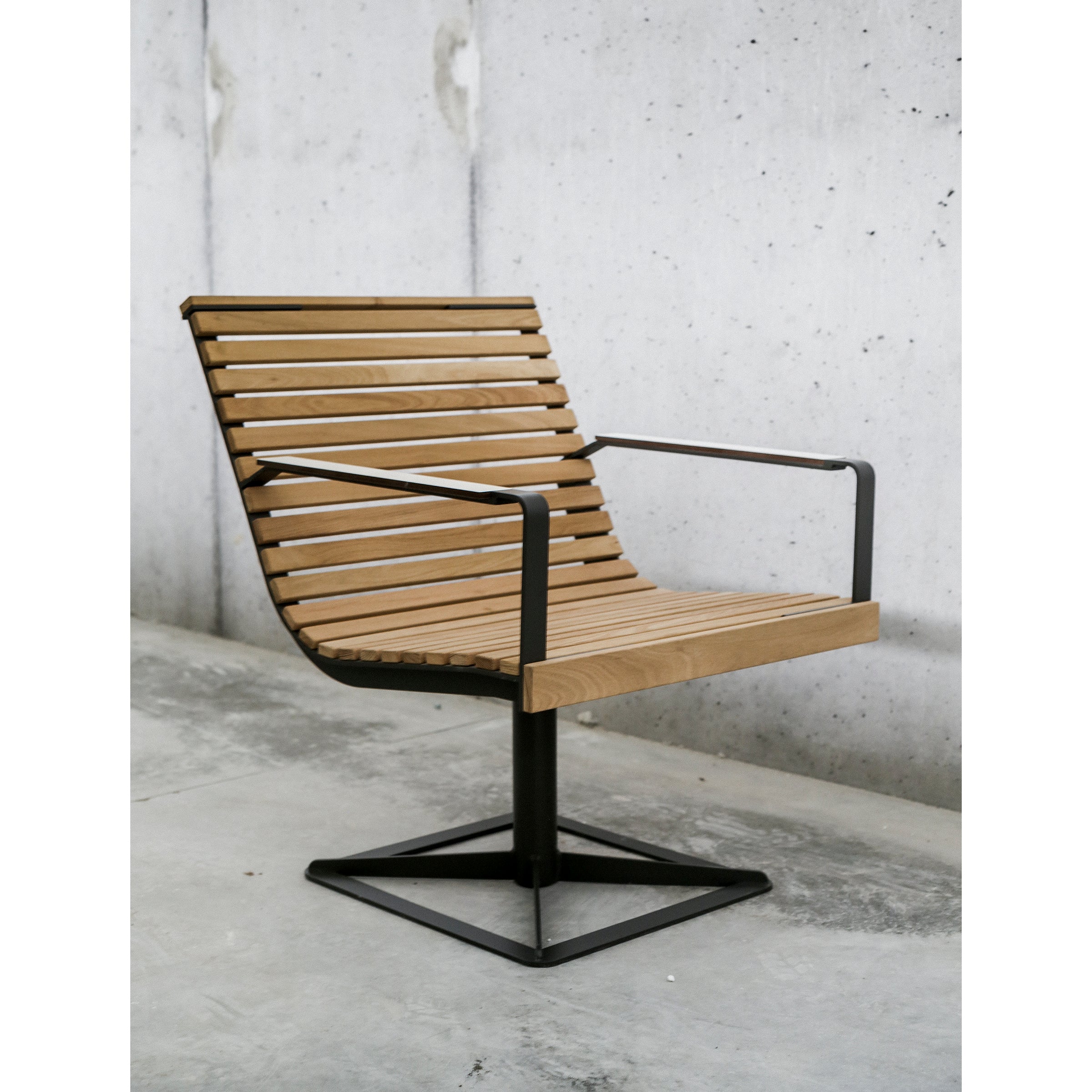 The Preva Armchair by Egoe boasts a sophisticated design with a wood slat look and metal frame, positioned on a concrete floor. With armrests and a sturdy square base, it embodies minimalist style against the plain wall, highlighting its enduring durability.