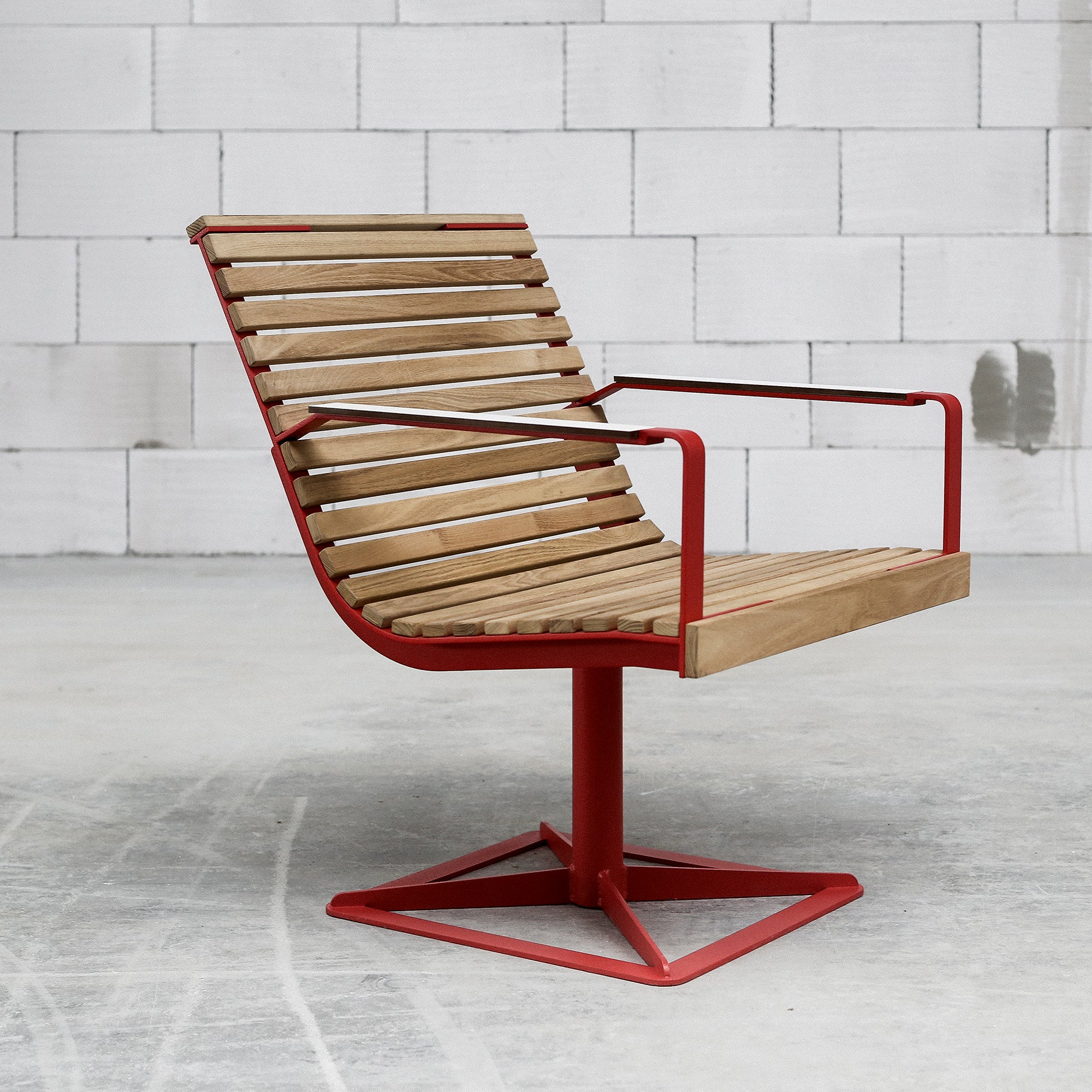 Introducing the Preva Armchair by Egoe: a sleek design with wooden slats, red metal armrests, and base. It sits on a concrete floor against a white cinder block wall, combining durability with an industrial minimalist look.
