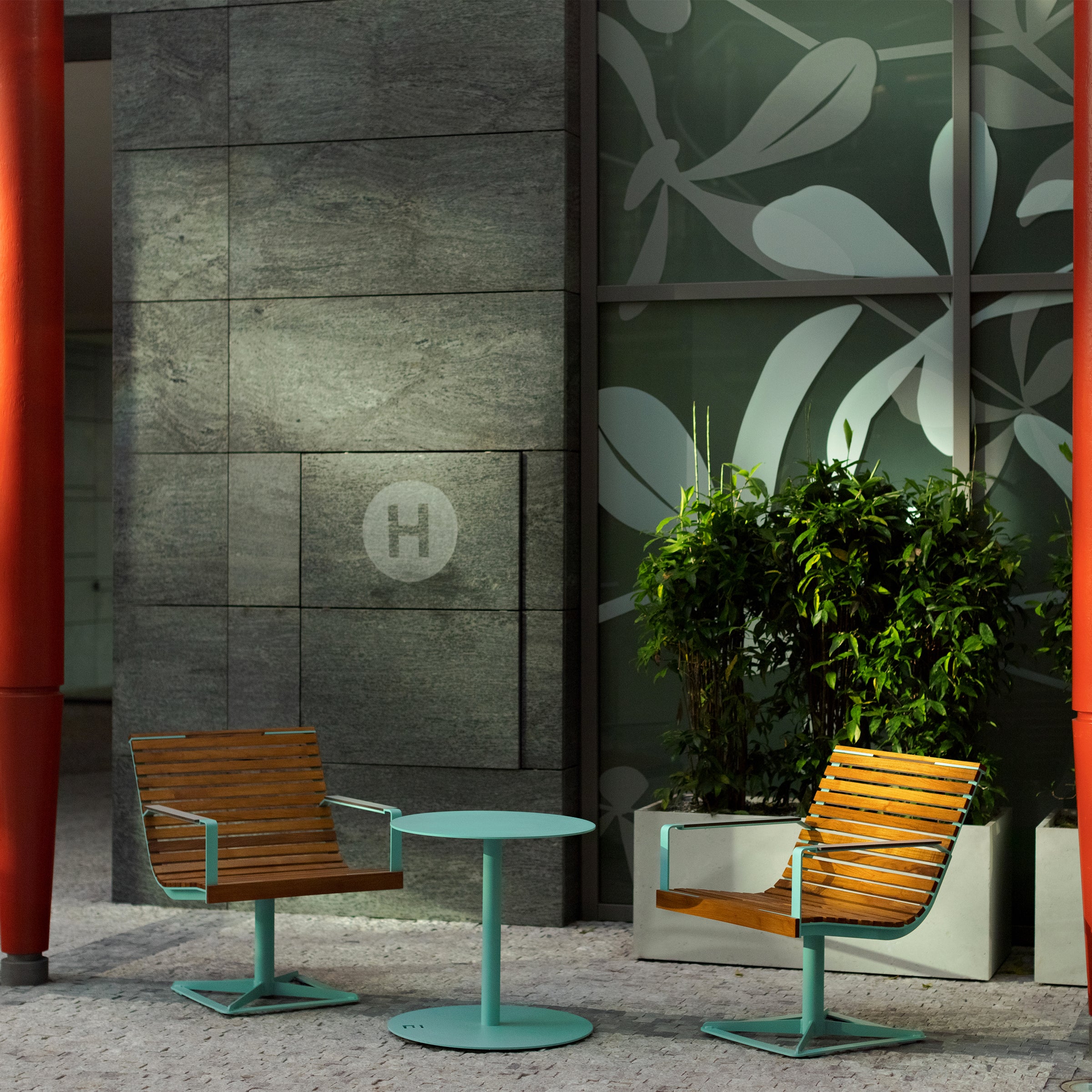 Two modern wooden chairs with blue metal frames accompany the minimalist Spulka Table by Egoe on a paved surface. A leafy plant stands against a wall adorned with grey stone tiles and frosted glass featuring abstract leaf designs.