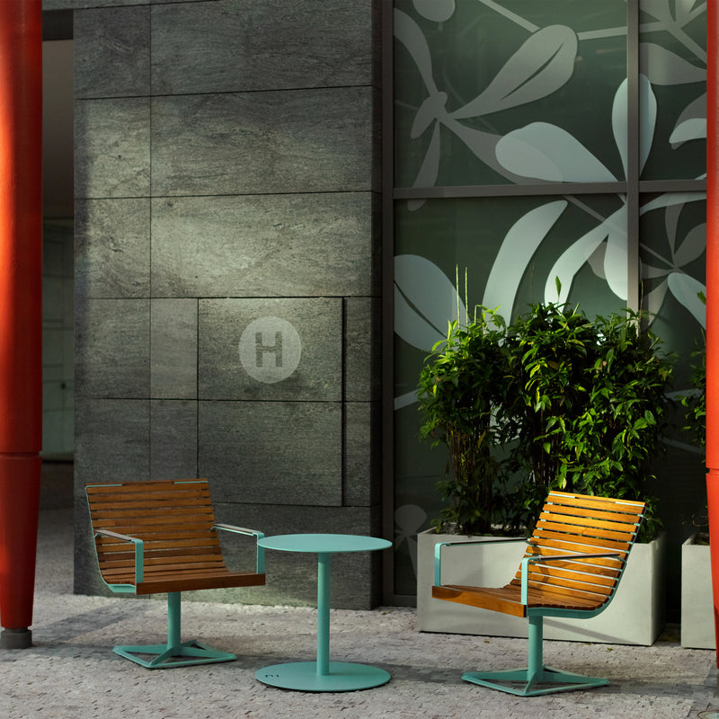 Two Preva Armchairs by Egoe, featuring teal metal frames and wood, along with a matching teal round table, are positioned against a dark stone wall adorned with a floral glass panel. A potted plant adds natures touch, while red pillars enhance the seating areas sophisticated design.