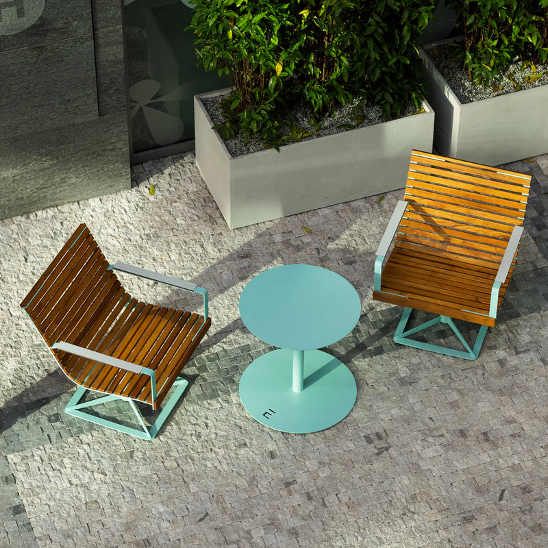 Two Preva Armchairs from Egoe, featuring wooden slats and metal frames, sit on either side of a small round turquoise table outdoors. These durable chairs add sophistication to the light-stone-paved space with large potted plants.