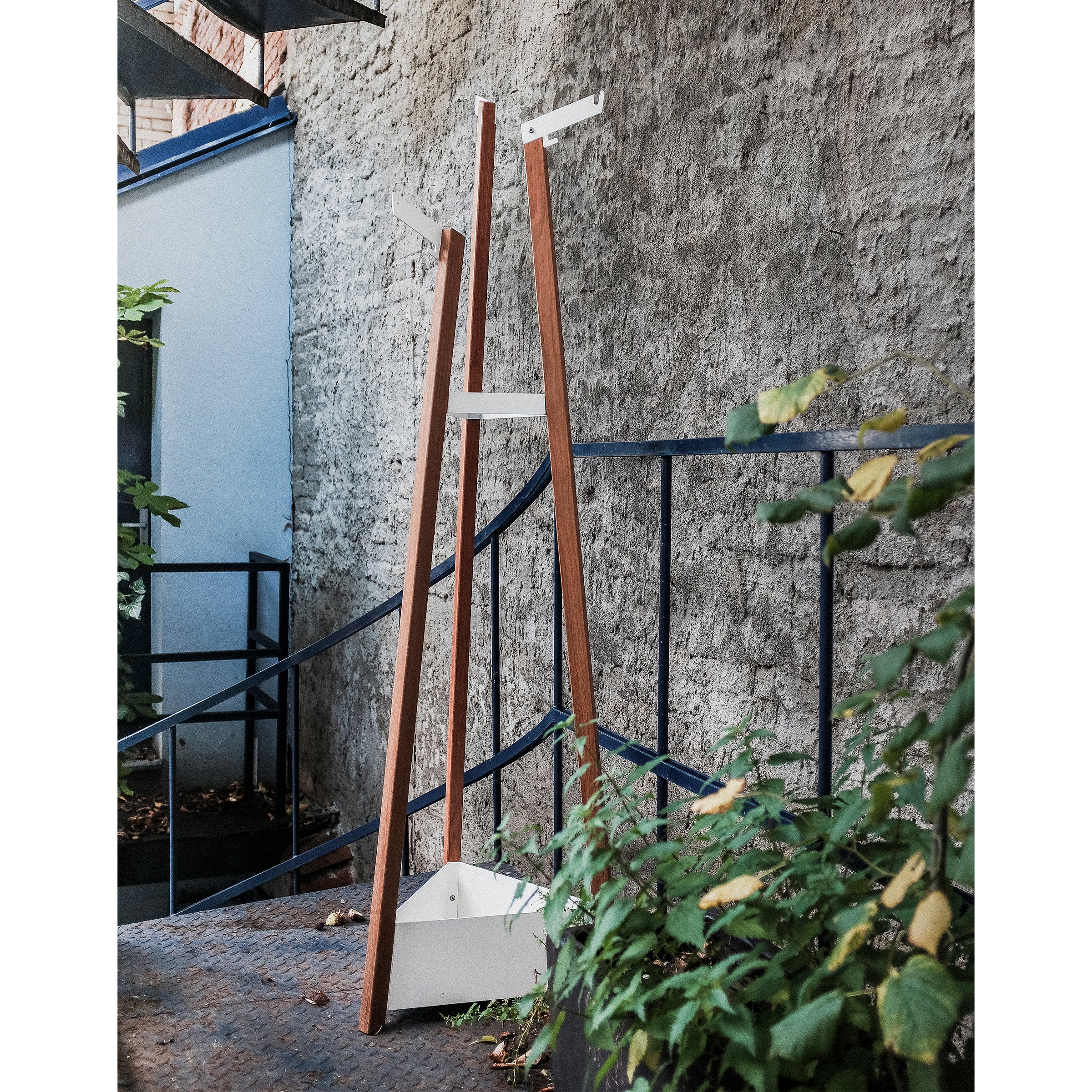 The Salto Coat Rack by Egoe, with wooden supports and a white base, stands on a metal staircase. Surrounded by concrete walls and lush greenery, it merges contemporary design with nature—a fitting addition to the stylish Salto collection.