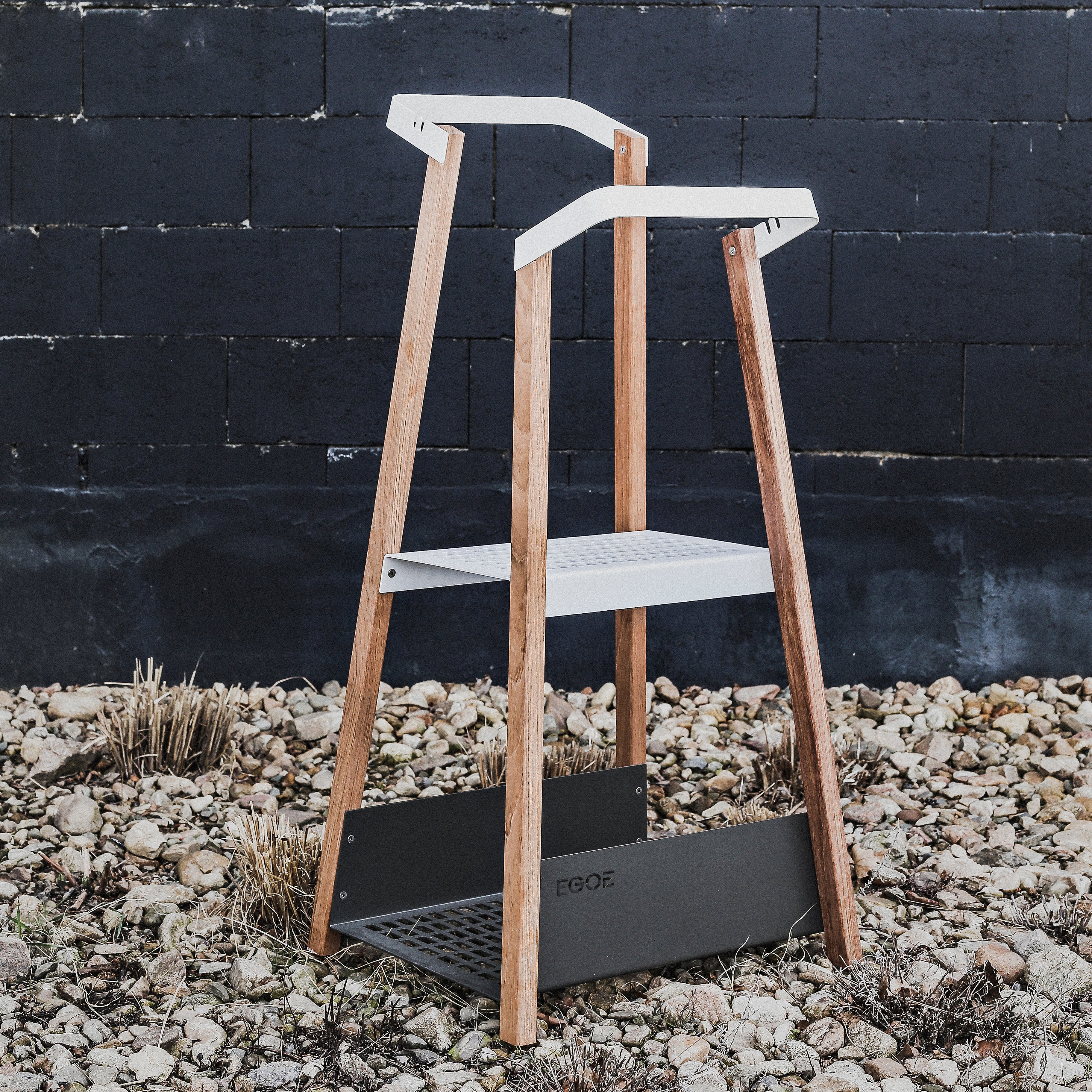 The Salto Coat Rack by Egoe, a modern blend of wood and metal, stands elegantly on rocks against a dark brick wall. It features a sleek two-tier design reminiscent of an outdoor plant stand with minimalist appeal.