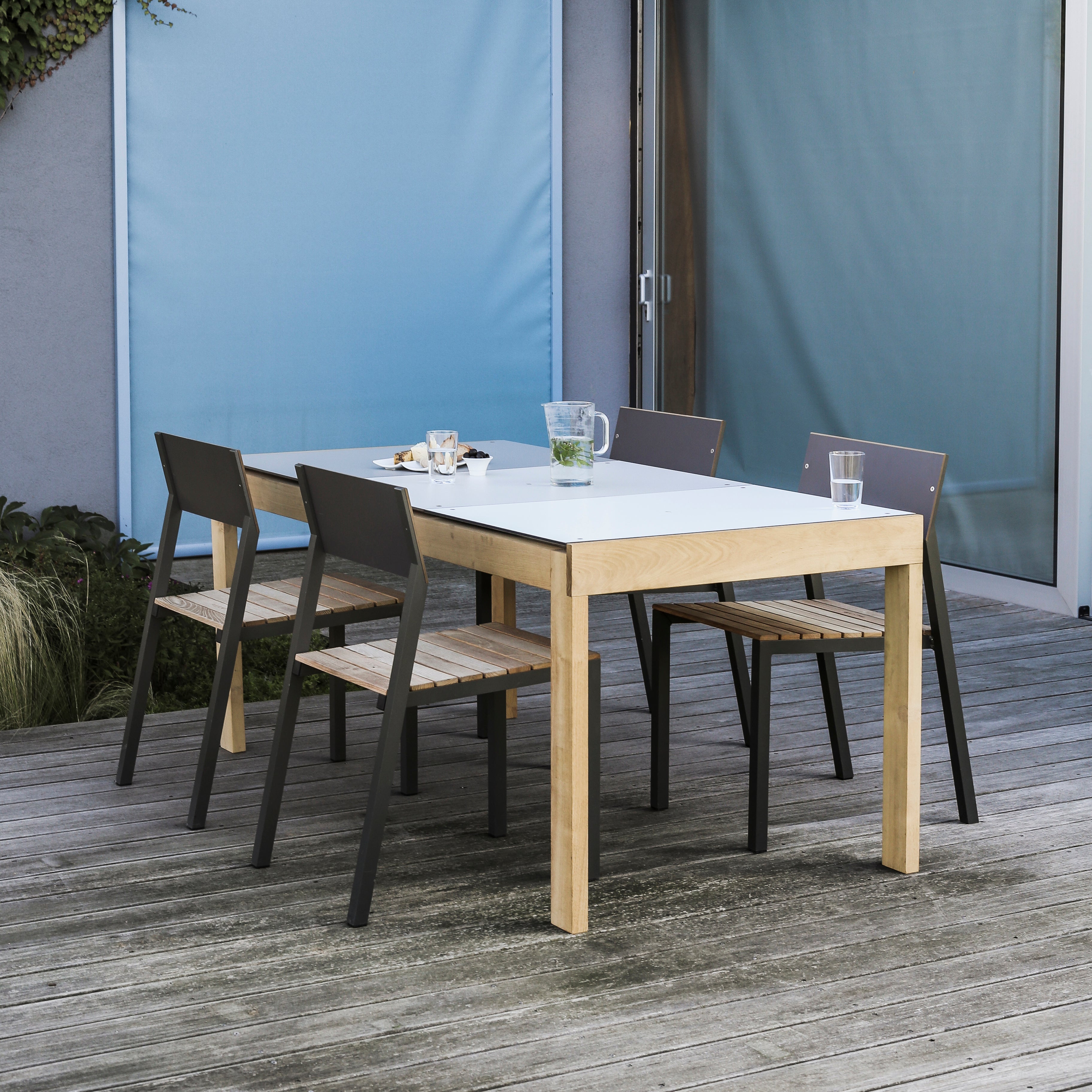 An outdoor dining area features an Egoe Sena Table with four chairs, two having dark backs. The light tropical wood table is adorned with a clear pitcher, two glasses, and a small plate. The setting enhances a wooden deck against a gray wall and blue panel backdrop.