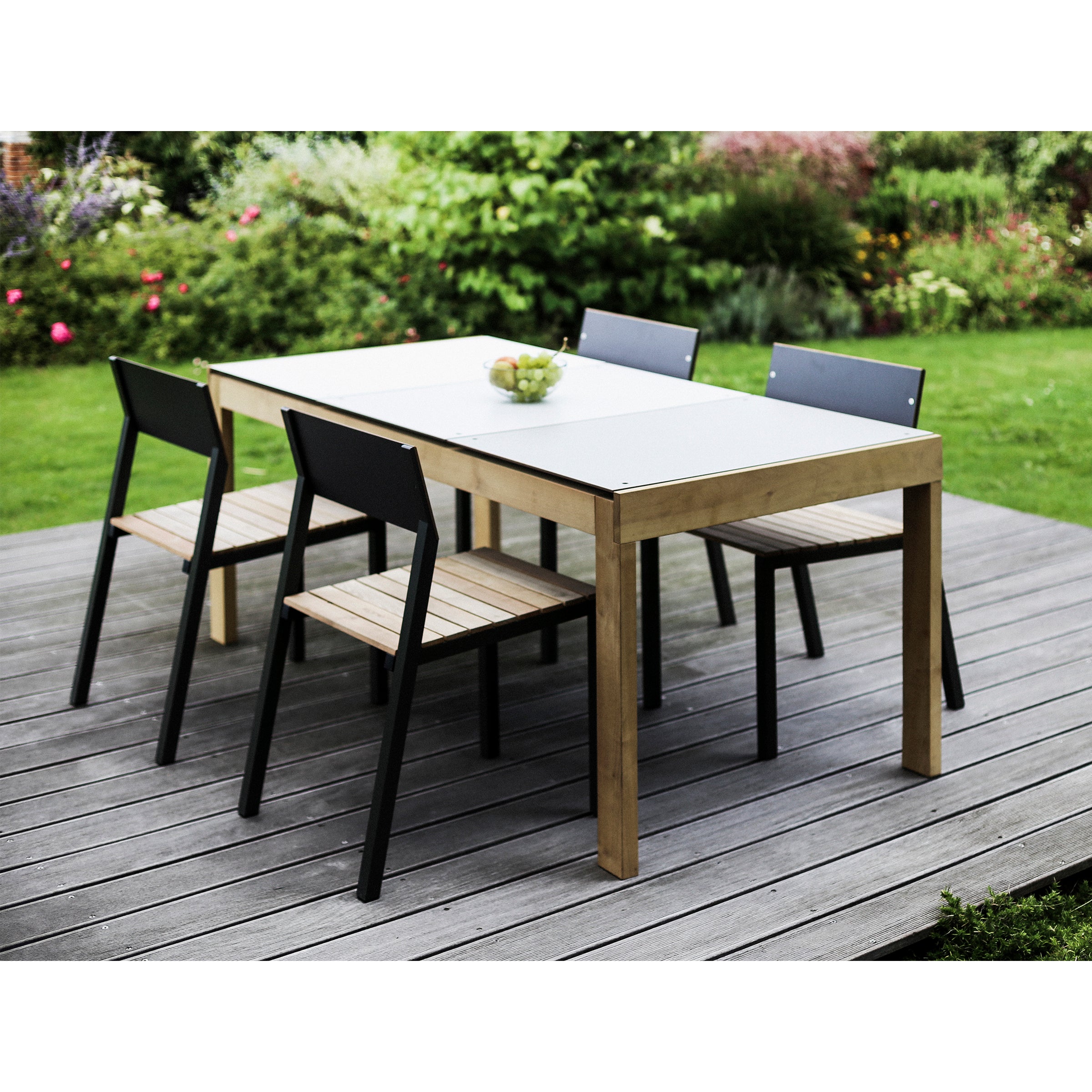 The outdoor dining set features the Sena Table by Egoe, crafted from tropical wood, with four black and wood chairs on a wooden deck. A bowl of apples graces the table amid a lush garden backdrop of grass and flowering plants.