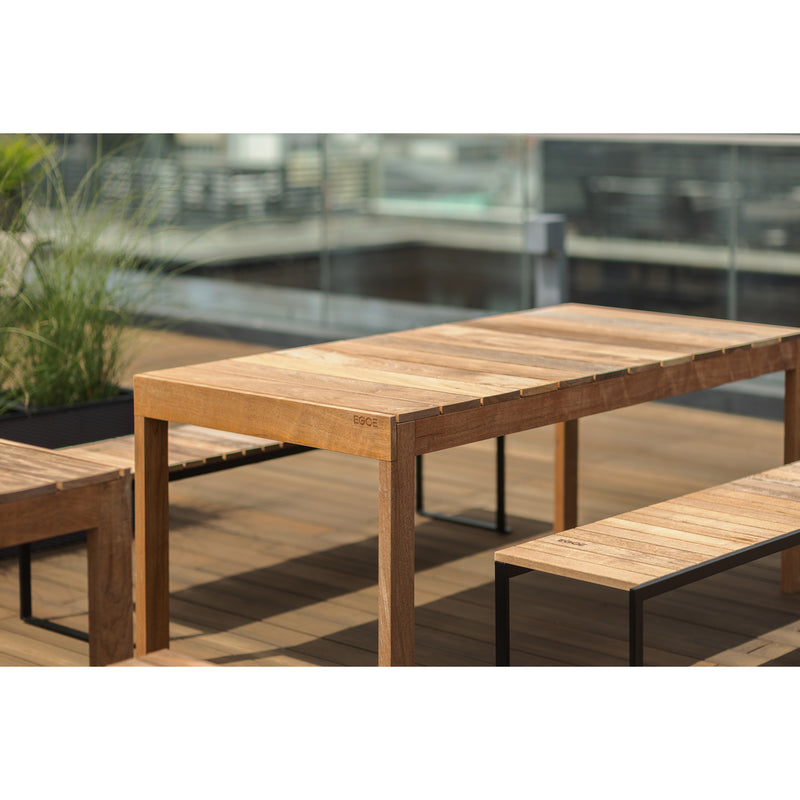 Egoes Sena Tables, crafted from tropical wood, grace the wooden deck alongside benches amid lush greenery in planters. A glass railing frames modern architectural views, blending nature with contemporary design.