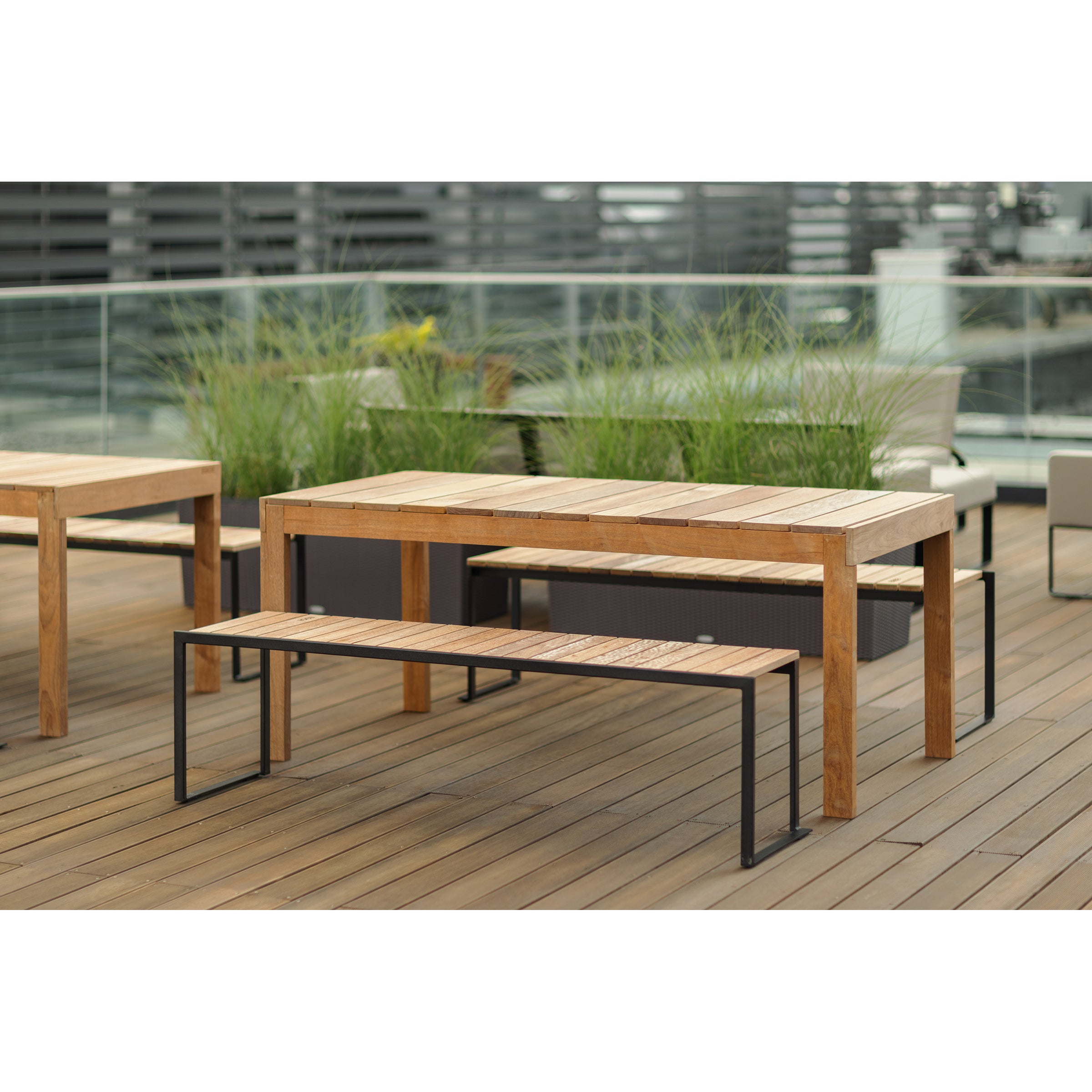 A wooden outdoor dining set made from tropical wood adorns a spacious deck, featuring a rectangular table and bench with sturdy metal frames. Tall plants in planters and sleek Sena tables by Egoe complement the glass railing, all set against a vibrant urban backdrop.