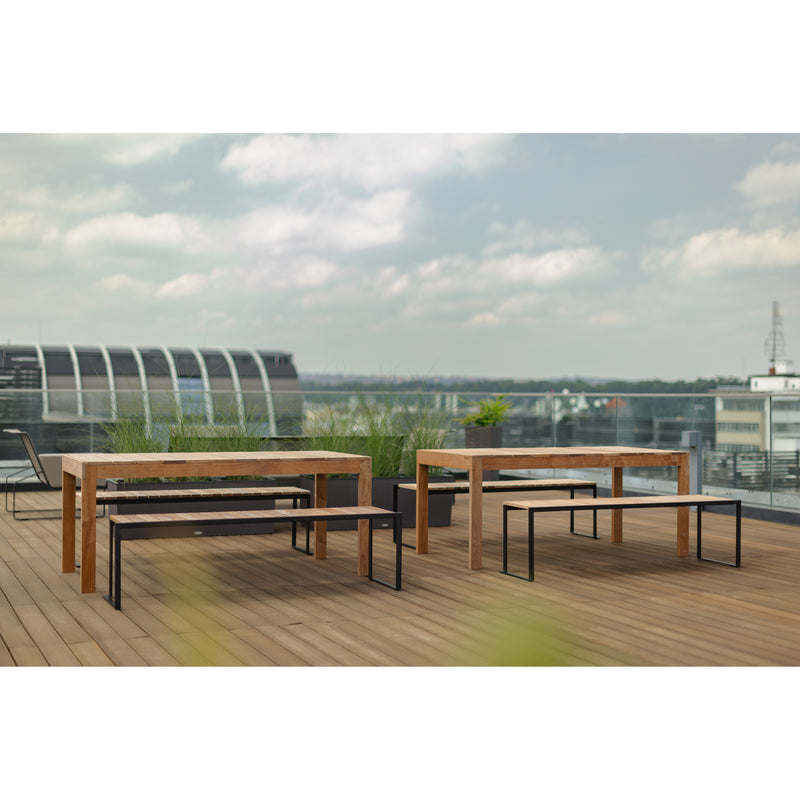 The rooftop deck is adorned with Egoes Sena tables and benches made from tropical wood, resting on a wooden floor. Planters with tall grass enhance the scene as a modern cityscape unfolds under a cloudy sky.