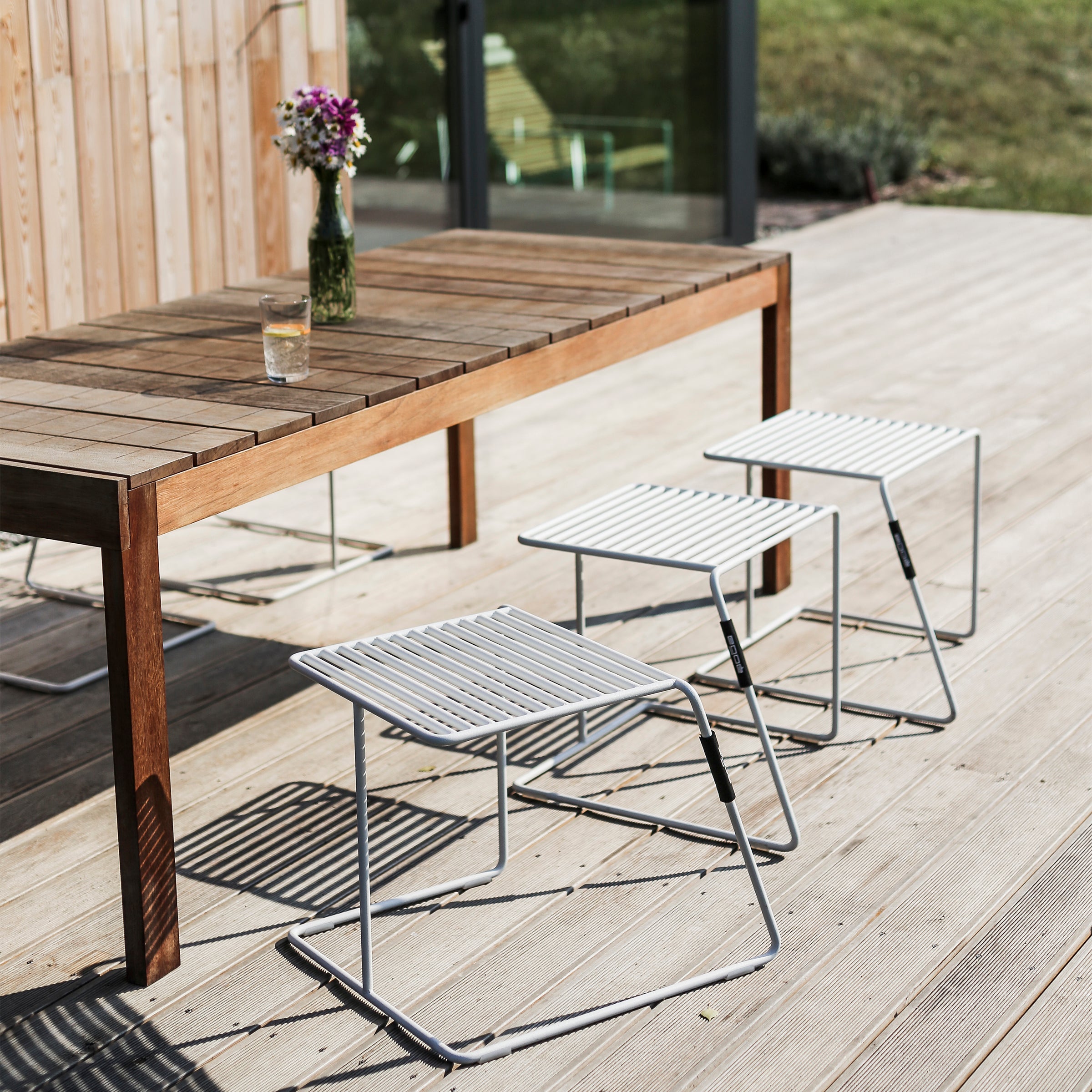 An Egoe Sena Table adorns the wooden deck, paired with three sleek metal stools, a vase of flowers, and a glass. Glass doors open to a grassy area, creating a peaceful, sunny vibe.