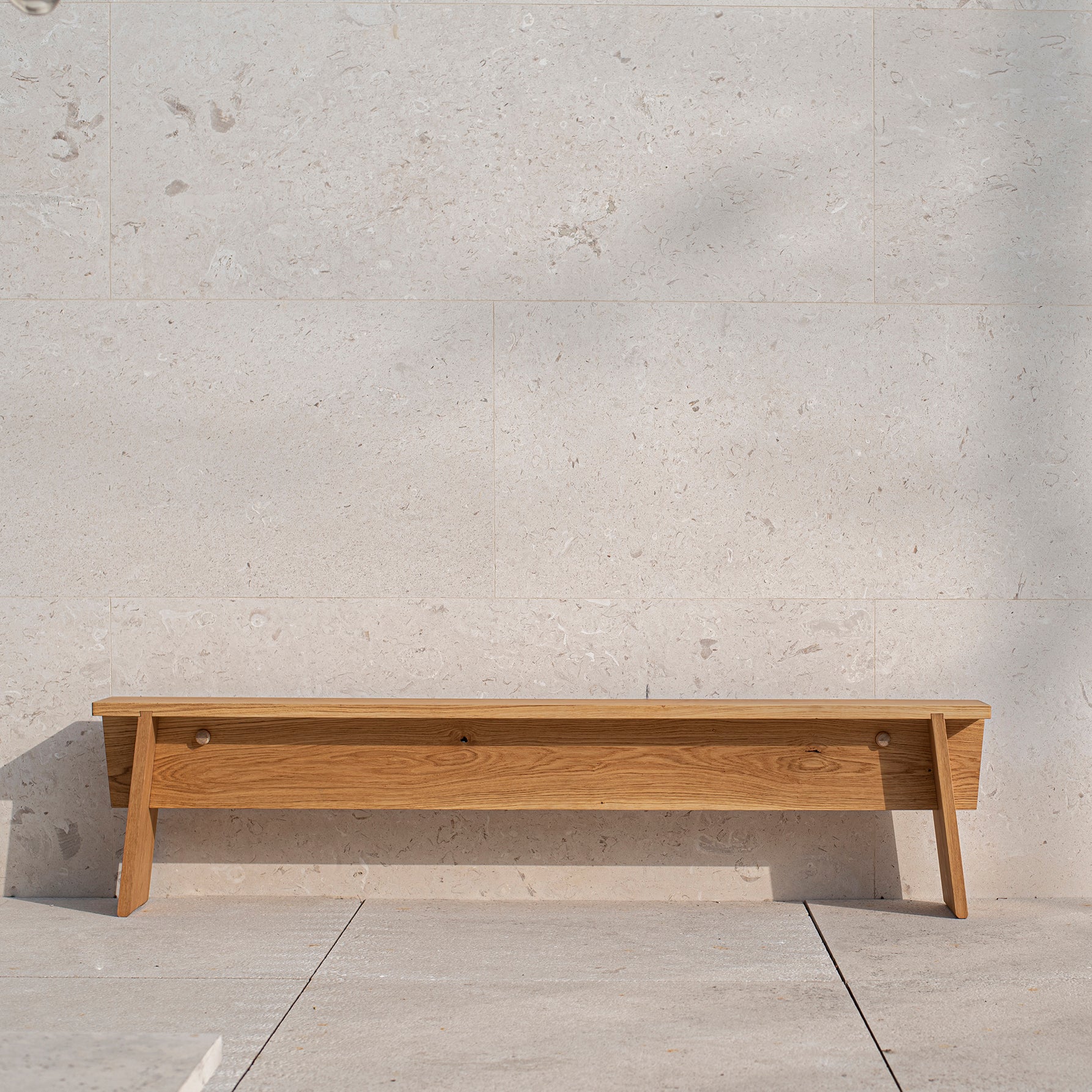 The Štok Bench by Egoe, crafted from thermal ash wood, complements a light beige stone wall on a stone-tiled floor with its minimalist design, enhancing the calm and orderly setting even when unoccupied.