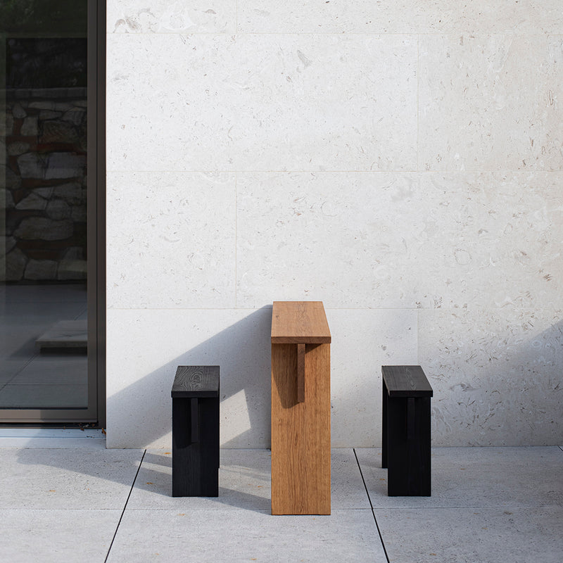 A serene minimalist outdoor setting showcases a small rectangular solid wood table with two Egoe Štok Stools in black, enhancing the elegance of a light-colored stone wall and glass door.
