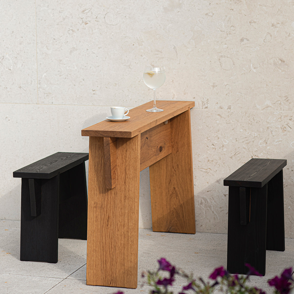 An outdoor setting showcases a tall Štok Bistro Table by Egoe, adorned with a glass of white wine and a coffee cup. Flanked by two black stools crafted from thermal ash wood, the scene is completed by a stone wall and purple flowers, creating a serene ambiance with timeless design.