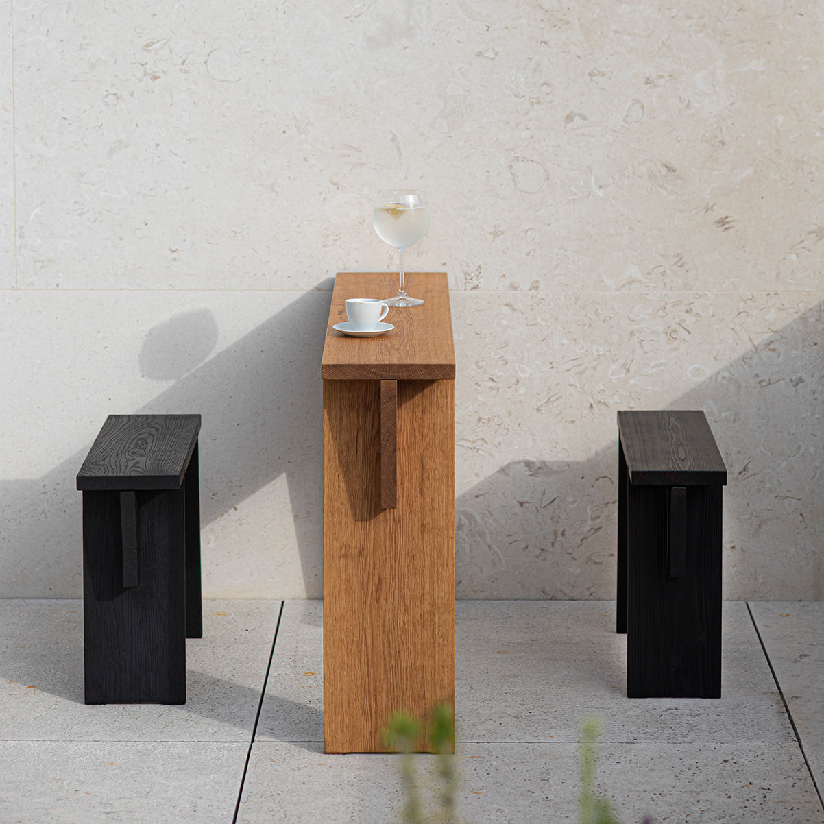A minimalist outdoor setup features the Štok Bistro Table by Egoé, crafted from thermal ash wood. It holds a glass of white wine and a coffee cup. Two low black benches, also by Egoé, flank the table against a light stone wall, highlighting its timeless design.