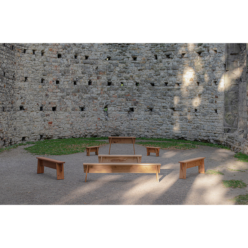A stone courtyard features several Štok Benches by Egoe, crafted from thermal ash wood and arranged in a semicircle. Sunlight paints shadows on the ground and walls, creating tranquility. A raised platform amidst grass and gravel completes this simple, timeless design.
