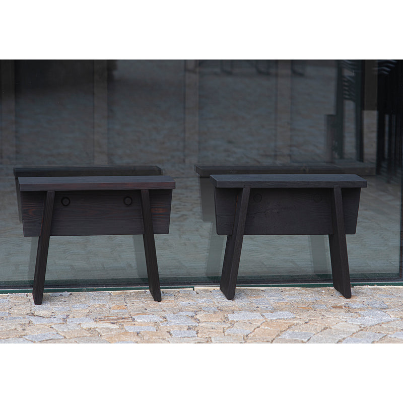 Two Štok Stools by Egoe, featuring a modern design with dark thermal ash and angular shapes, stand on a stone-tiled surface in front of glass doors.