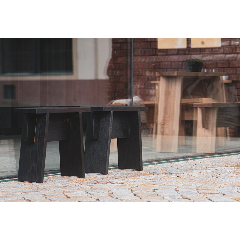 Two Štok Stools by Egoe, with a minimalist black wooden design, sit outside a glass window. Inside, more solid wood stools and a table are visible against the cobblestone floor and red brick wall backdrop.