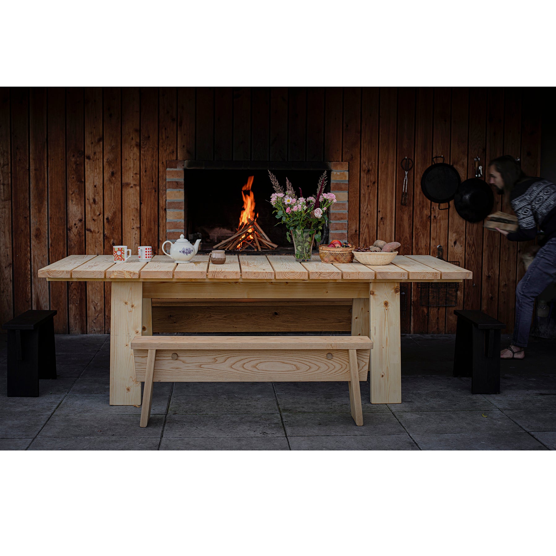 A cozy wooden table with mugs and bread in front of a lit fireplace. Someone is using an easy-to-assemble shelf on the right. Rustic walls and the Štok Bench by Egoe add to the inviting atmosphere.