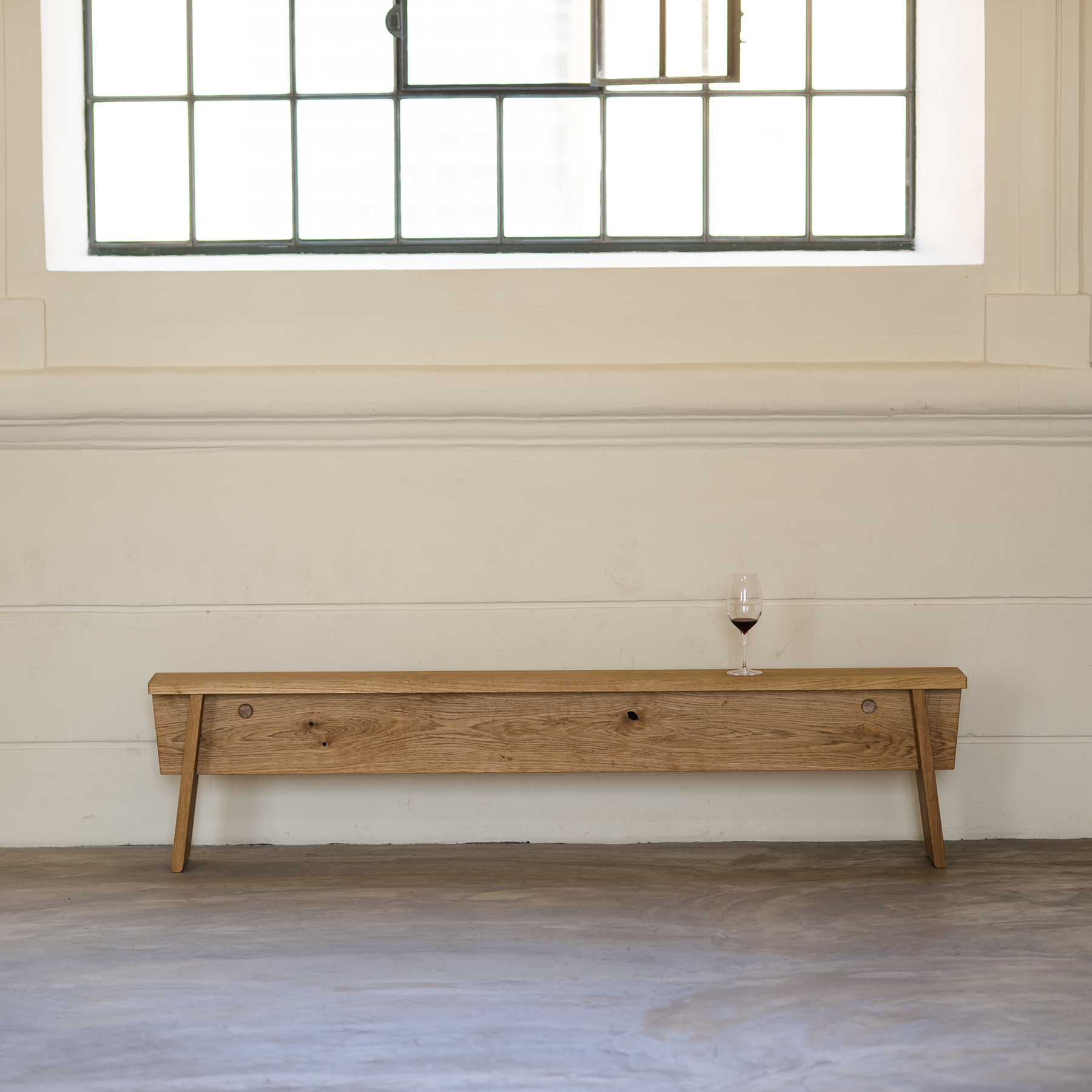 The Štok Bench by Egoe, a minimalist piece crafted from thermal ash wood, elegantly holds a glass of red wine in front of a cream wall. A large grid-pane window filters natural light into the space, highlighting its timeless, easy-to-assemble design.