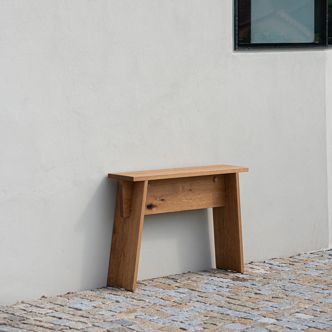 The Štok Stool by Egoe, in solid wood with a natural finish, is set against a white wall on gray and brown stones. In the upper right corner, a dark window frame highlights the stools simplicity.