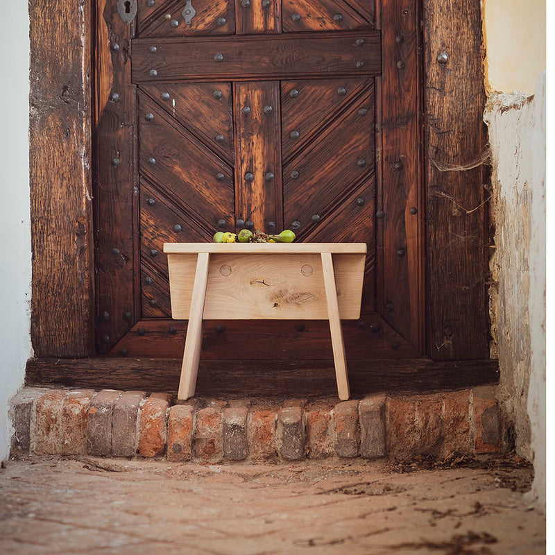 The Štok Stool by Egoe, made from solid wood, sits in front of an ornate wooden door with a geometric pattern and dark metal accents. Scattered small green apples enhance the old-world vintage charm with simple design elegance.