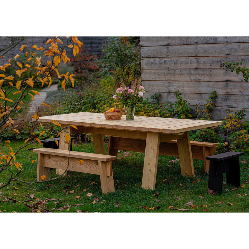 A timeless design defines this picnic table set in a garden with autumn foliage. A vase of flowers and a wooden bowl with apples rest on the table. Two benches flank either side, crafted from Egoes Štok Bench collection in thermal ash wood, plus a small black stool on one end.