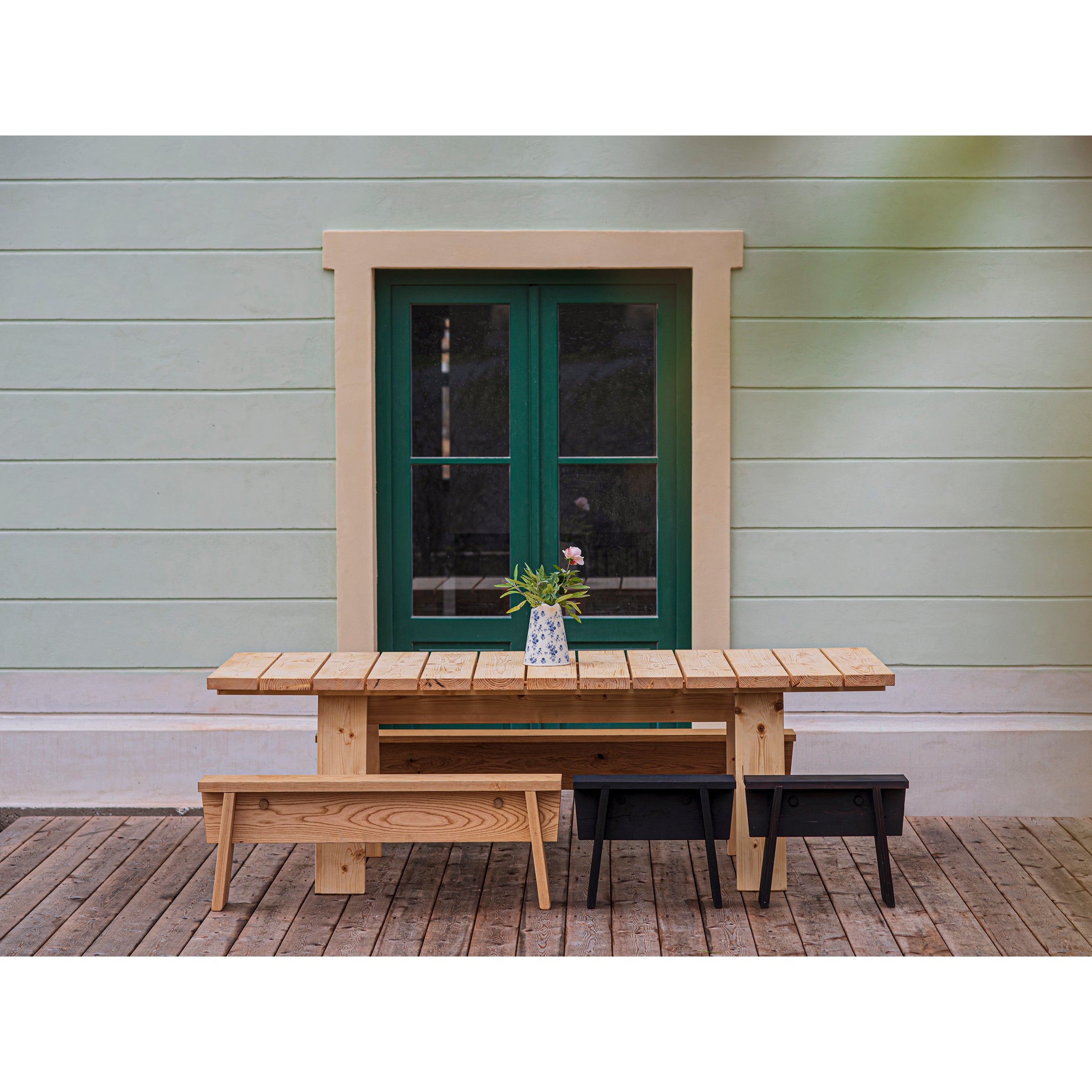 A timeless scene features the Egoe Štok Bench and table, crafted from thermal ash wood, placed in front of a house with pastel green walls and a green-framed window. A vase of flowers adorns the table, set on an easy-to-assemble plank floor.