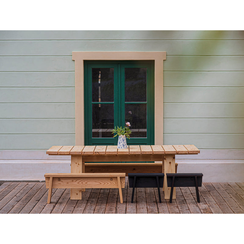 A timeless scene features the Egoe Štok Bench and table, crafted from thermal ash wood, placed in front of a house with pastel green walls and a green-framed window. A vase of flowers adorns the table, set on an easy-to-assemble plank floor.