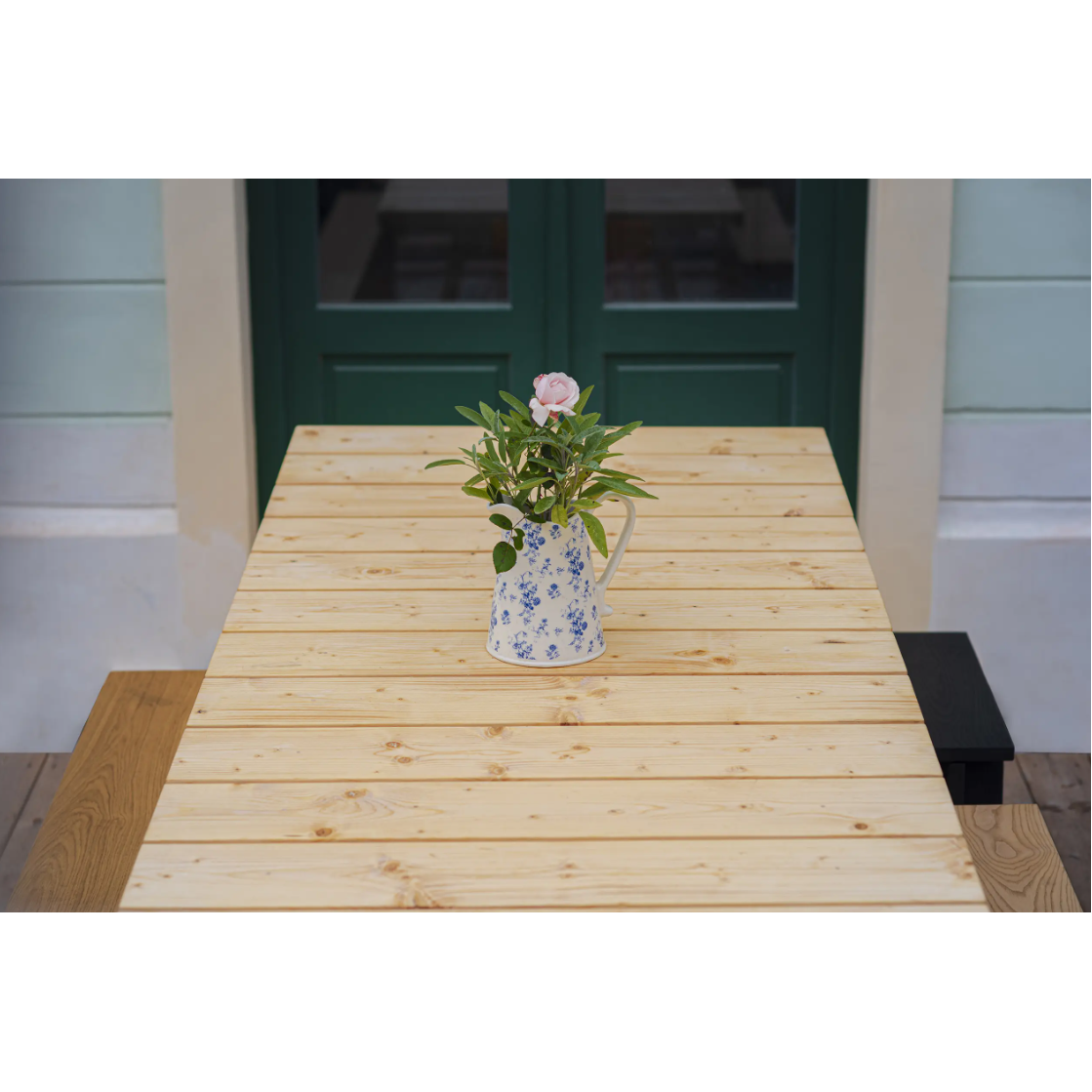The Štok Dining Table from Egoes timeless collection features a floral-patterned pitcher with pink roses and greenery. Green French doors against light walls enhance the classic aesthetic.