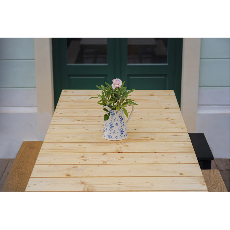 The Štok Dining Table from Egoes timeless collection features a floral-patterned pitcher with pink roses and greenery. Green French doors against light walls enhance the classic aesthetic.