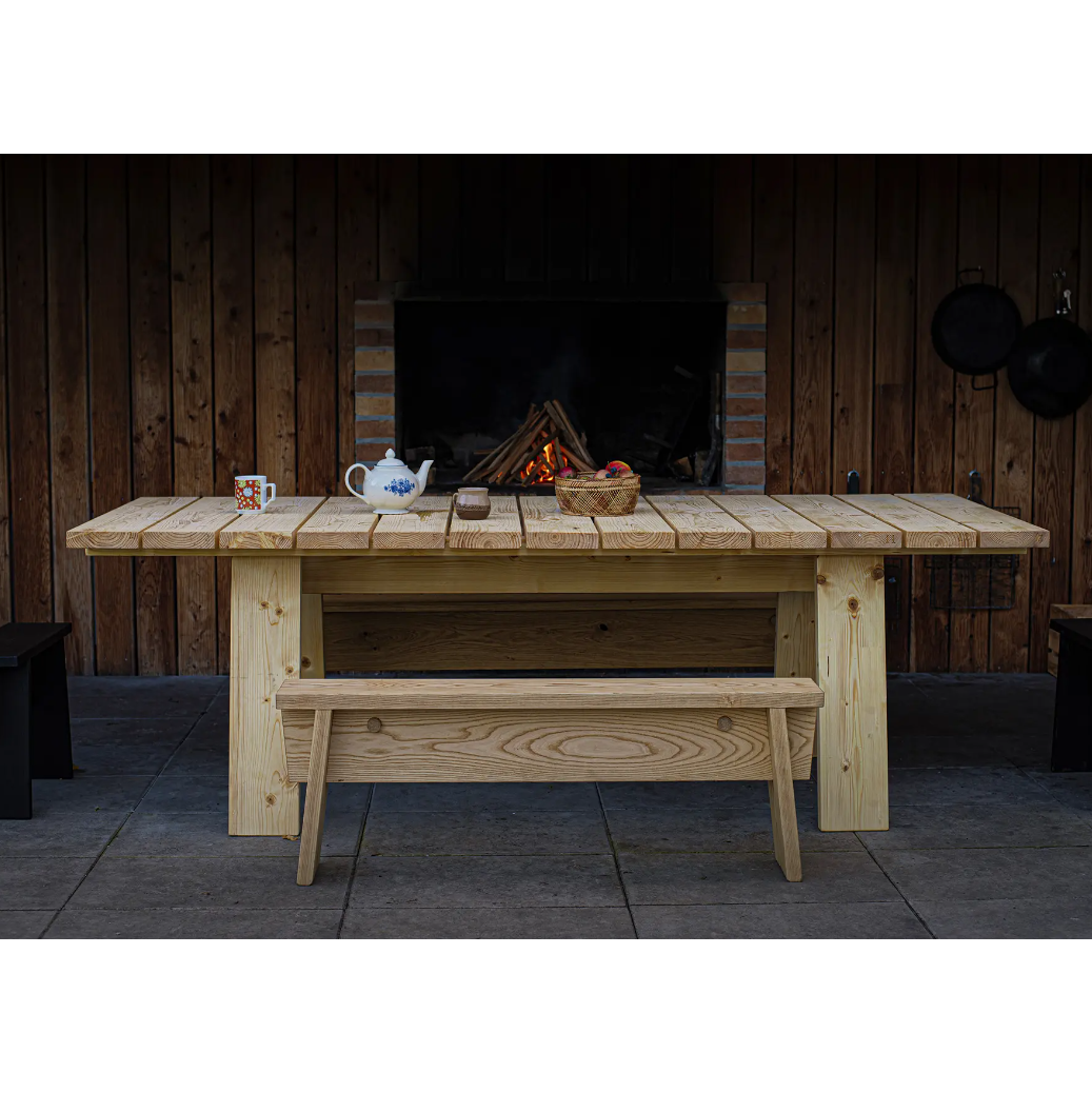 The Štok Dining Table by Egoe, alongside a bench from our timeless wood collection, complements a patio in front of a rustic fireplace with wooden walls. On the table are a teapot, cup, and basket of apples. Hanging pans enhance this charming ambiance.
