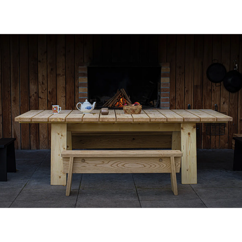 The Štok Dining Table by Egoe, alongside a bench from our timeless wood collection, complements a patio in front of a rustic fireplace with wooden walls. On the table are a teapot, cup, and basket of apples. Hanging pans enhance this charming ambiance.
