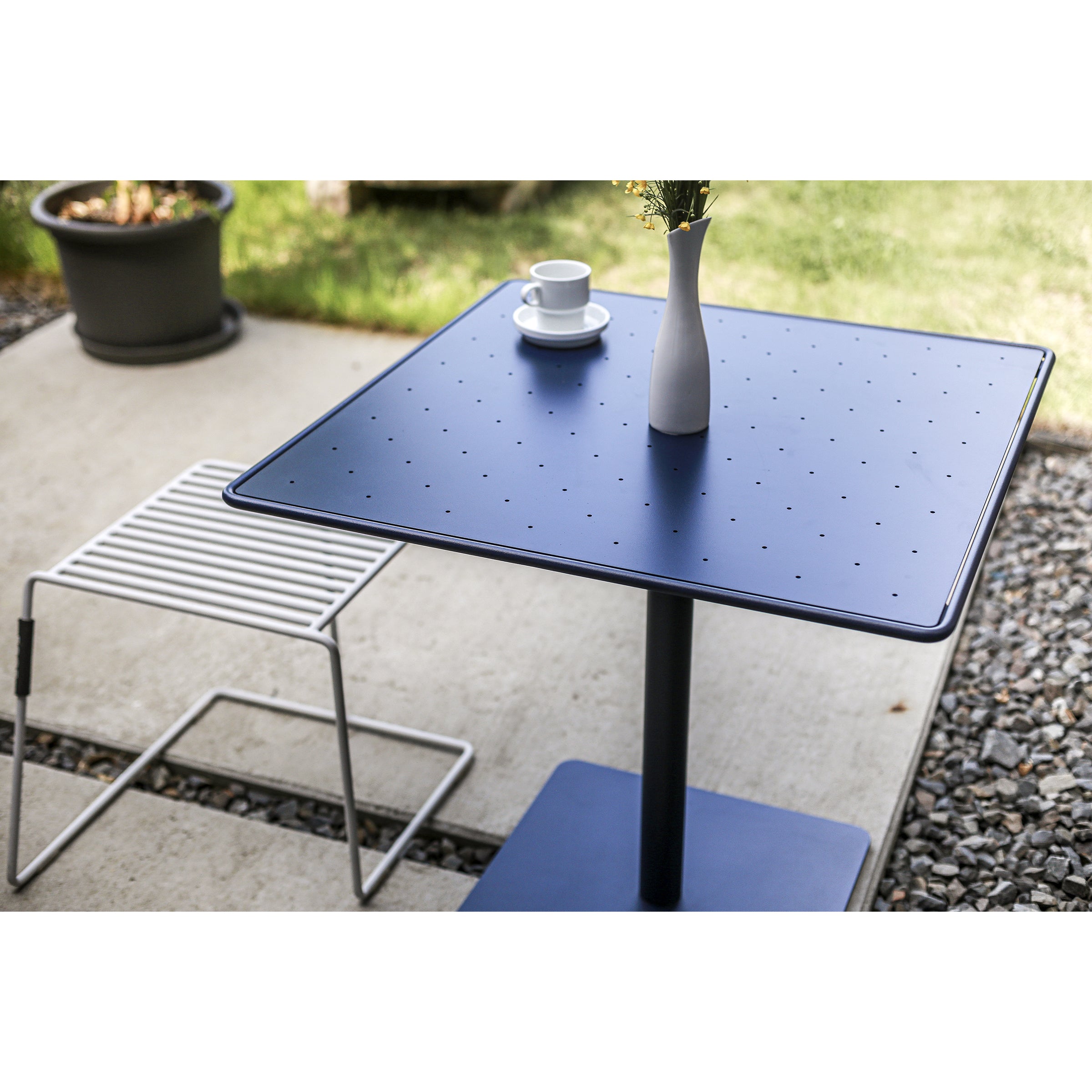 A Tina Table by Egoe sets a contemporary patio scene with its blue square design. Complemented by a small white vase of flowers, cup and saucer, and a white wire-frame chair, the arrangement on concrete amid potted plants and gravel reflects an optimistic color palette.