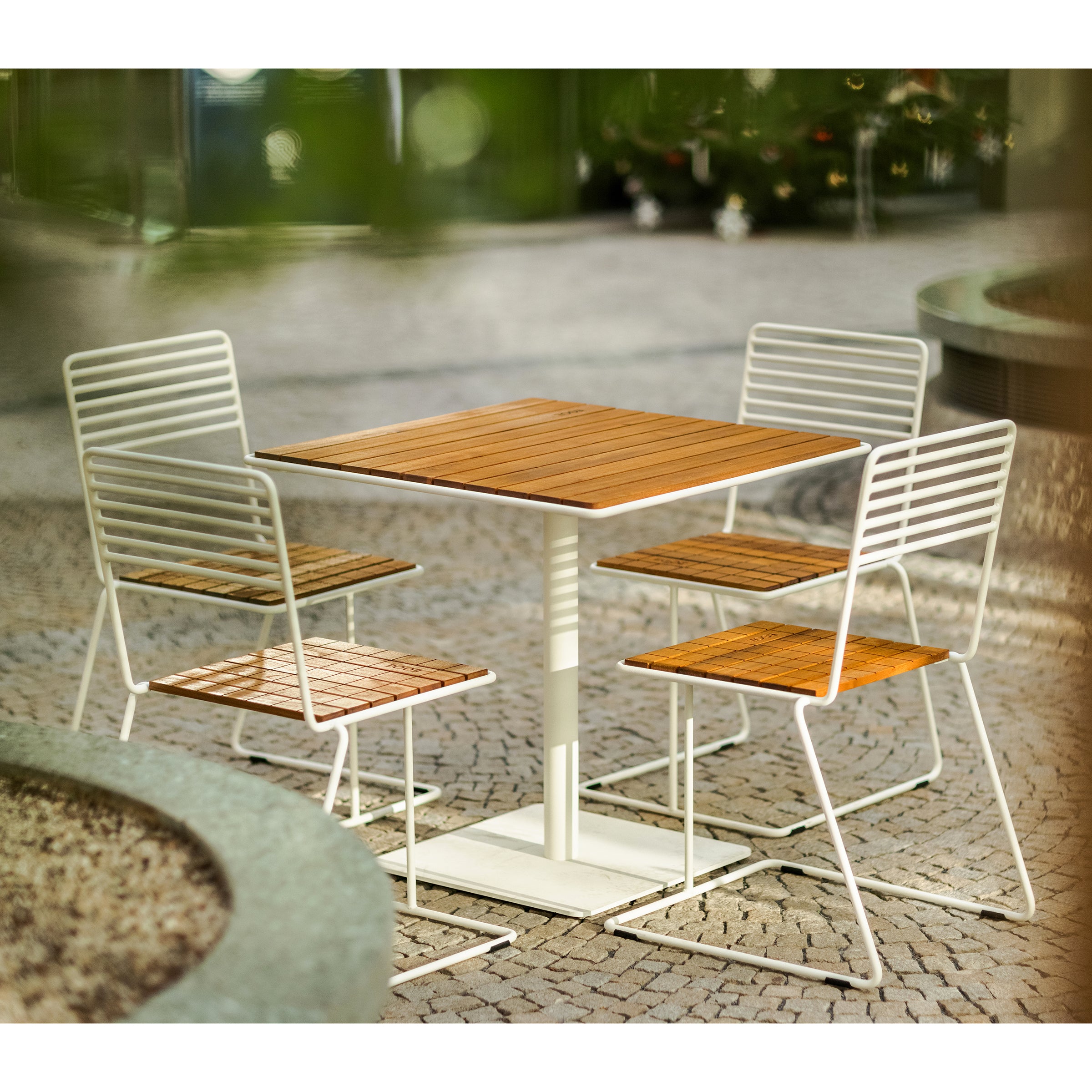 The Egoe Tina Table and four matching chairs create a serene patio setting. The chairs white metal frames add visual lightness, while their wooden seats and backrests beautifully complement the lush greenery.