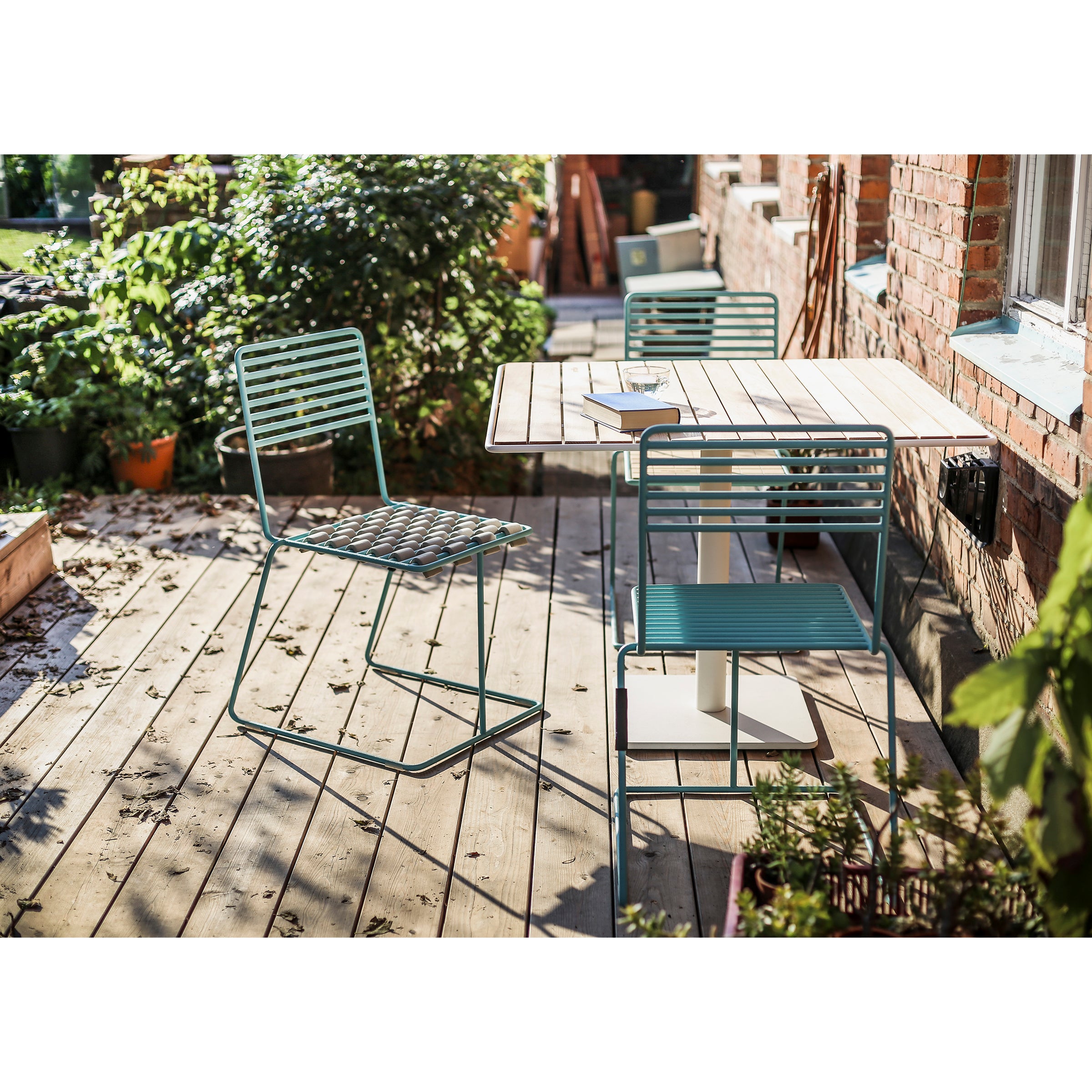 A patio with a wooden deck features an optimistic color scheme, highlighting a green metal chair and matching bench around Egoes Tina Table. Sunlight creates pleasing shadows, while potted plants near the brick building add visual lightness to the space.