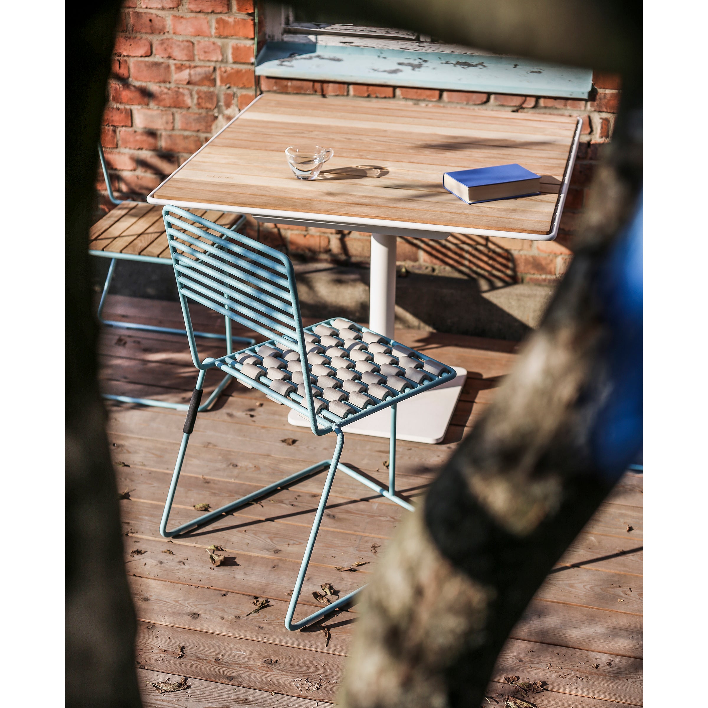 A wooden patio table with a powder-coated finish holds glasses and a closed blue book. Beside it is the Tina Chair by Egoe, featuring a patterned cushion. Sunlight filters through tree branches, casting gentle shadows in this tranquil outdoor setting.